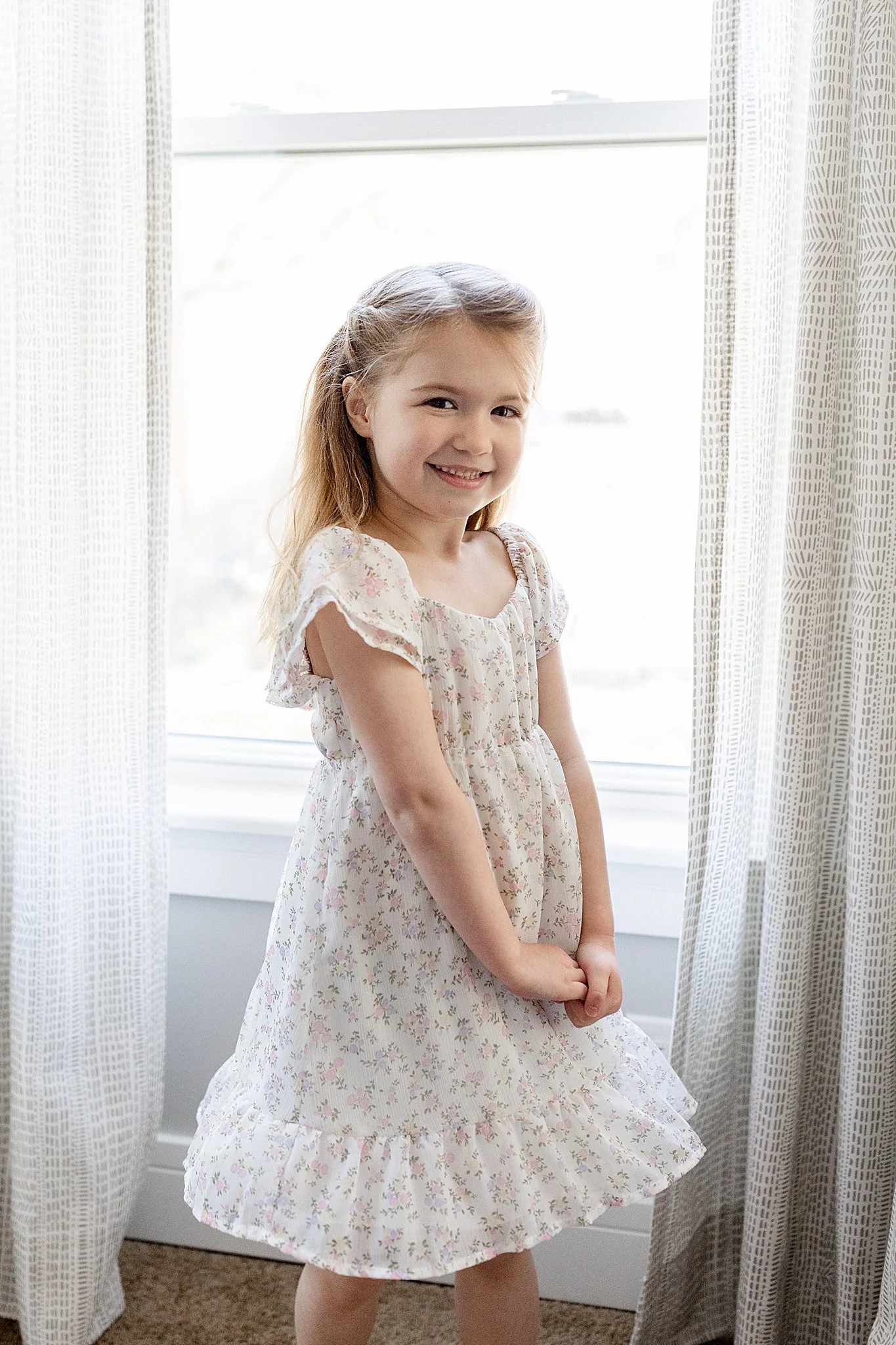 A young girl with blonde hair standing in front of a window with white textured curtains, smiling and holding her hands together in front of her, wearing a floral dress with a lifestyle photographer Naperville.