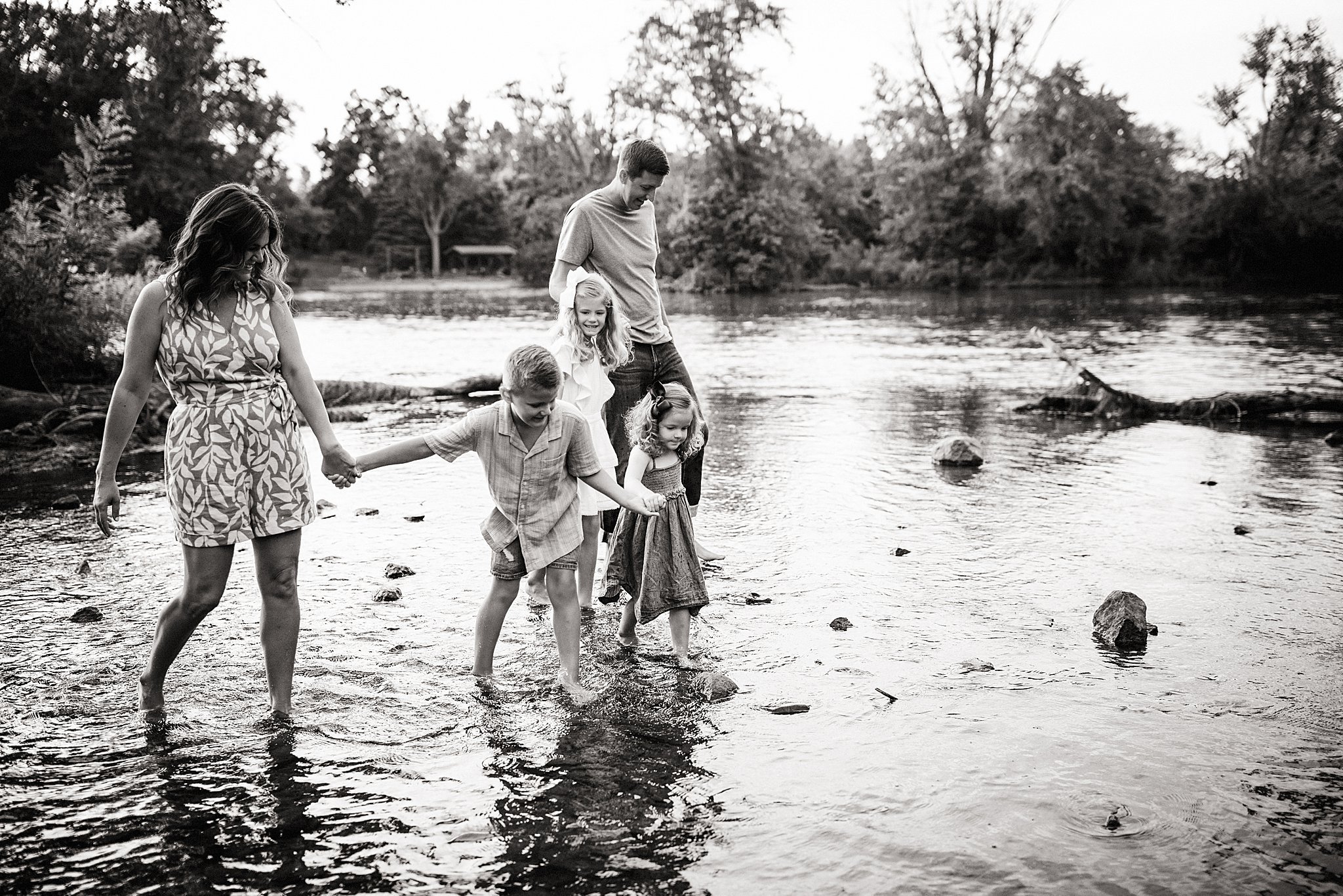 A family of five, including two adults and three children, wading through a river holding hands, surrounded by trees and nature in black and white during a creek mini session with Ally and B Photography in Oswego, IL.
