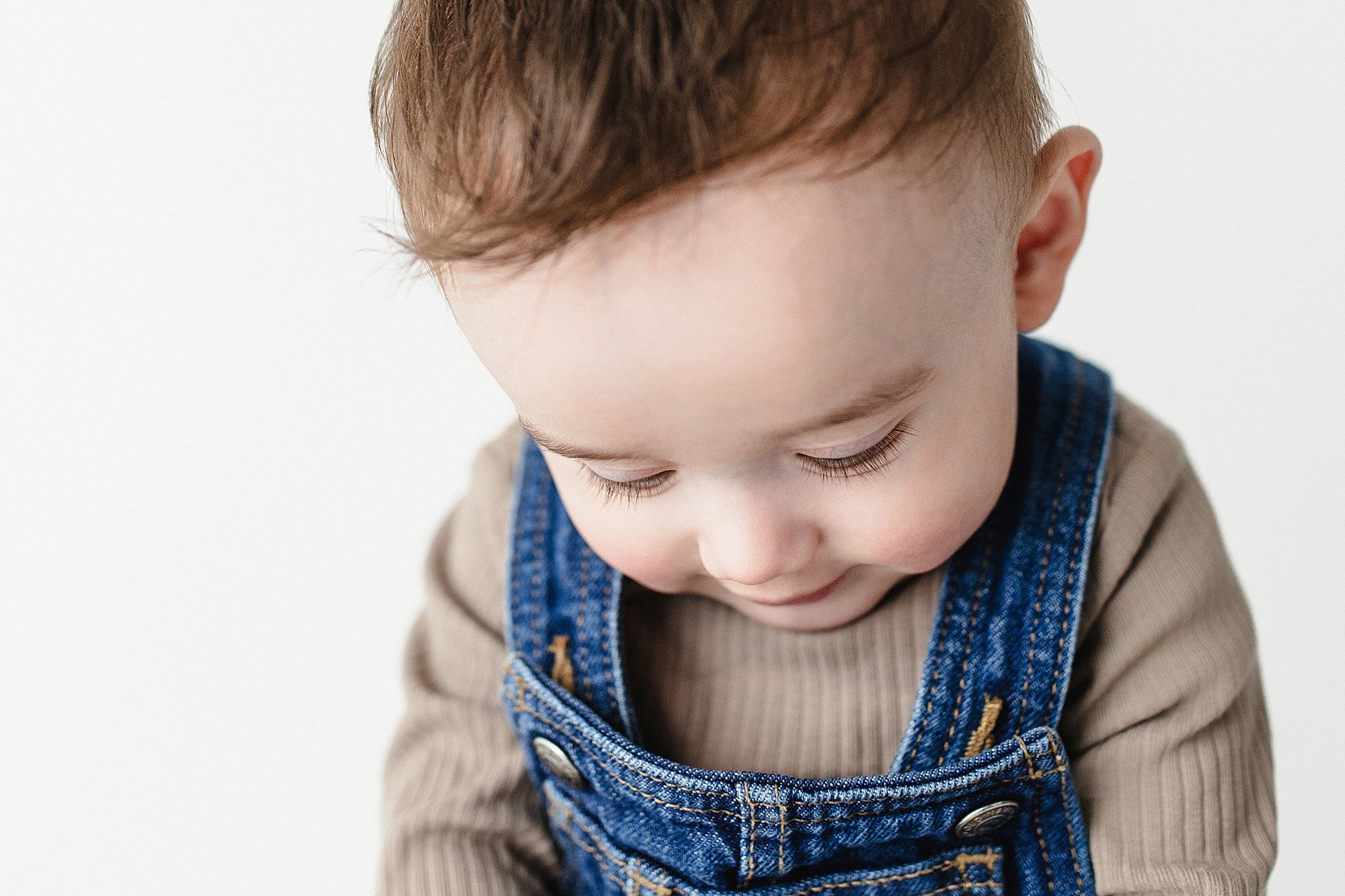 Close-up of a young boy with brown hair looking down, wearing a beige long-sleeve shirt and blue denim overalls, against a white background for infant photographer near me.
