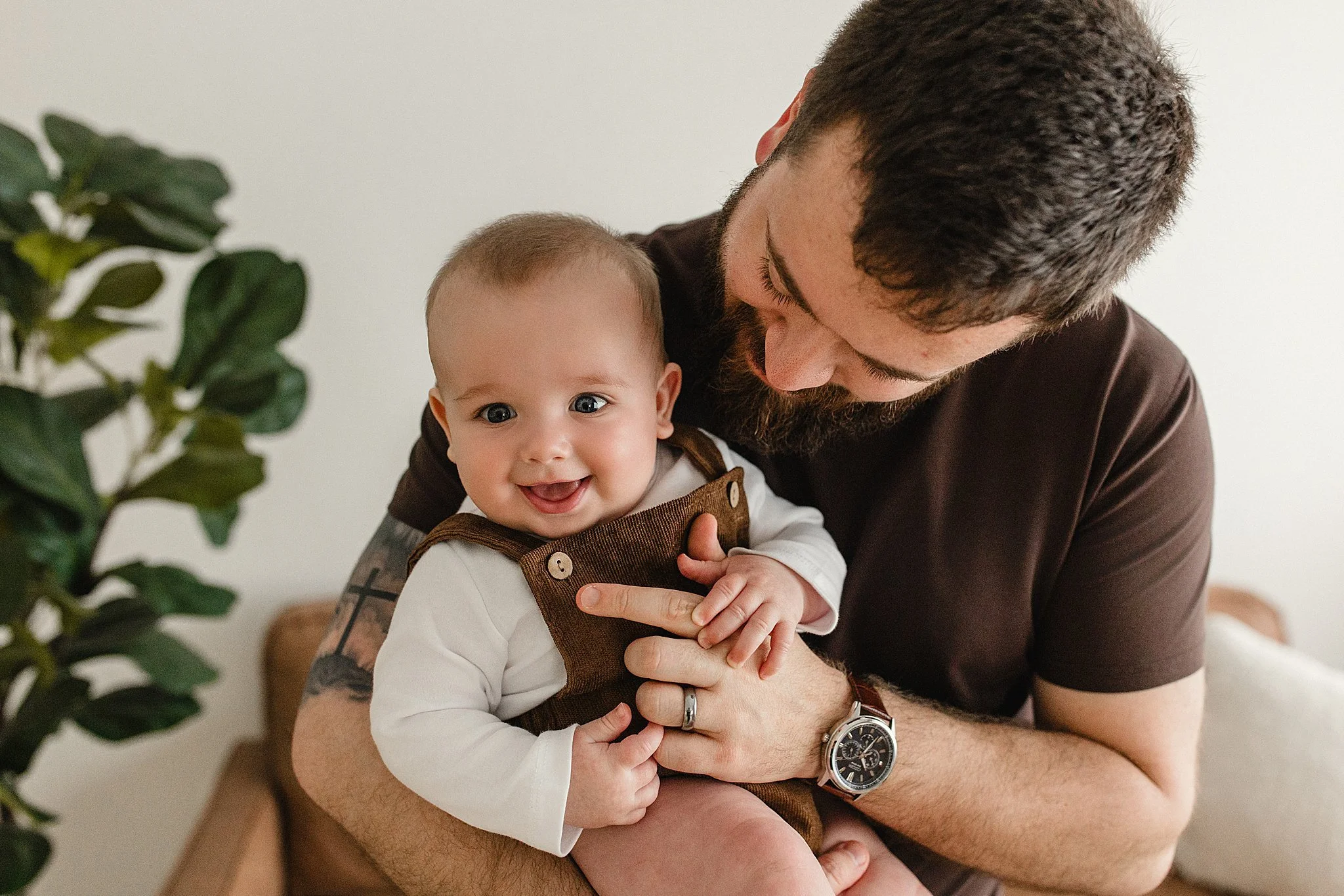 A man with a beard and short hair holding a laughing baby girl with blue eyes and light skin, sitting on the man's lap indoors with a white wall and green plant in the background during family photos near me.