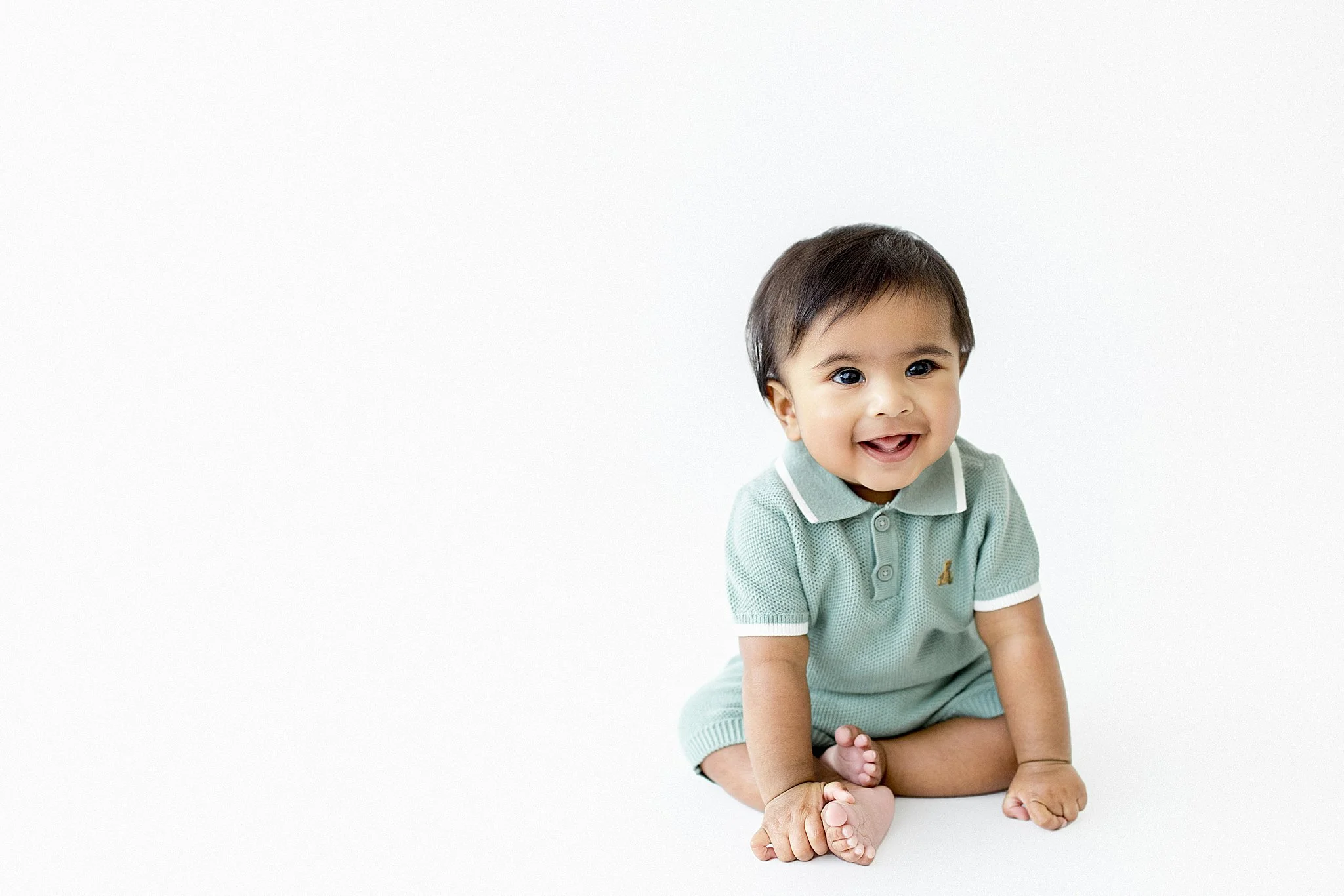 A smiling baby with dark hair, wearing a mint green polo shirt and shorts, sitting on the floor against a white background during a milestone session with Ally and B Photography.