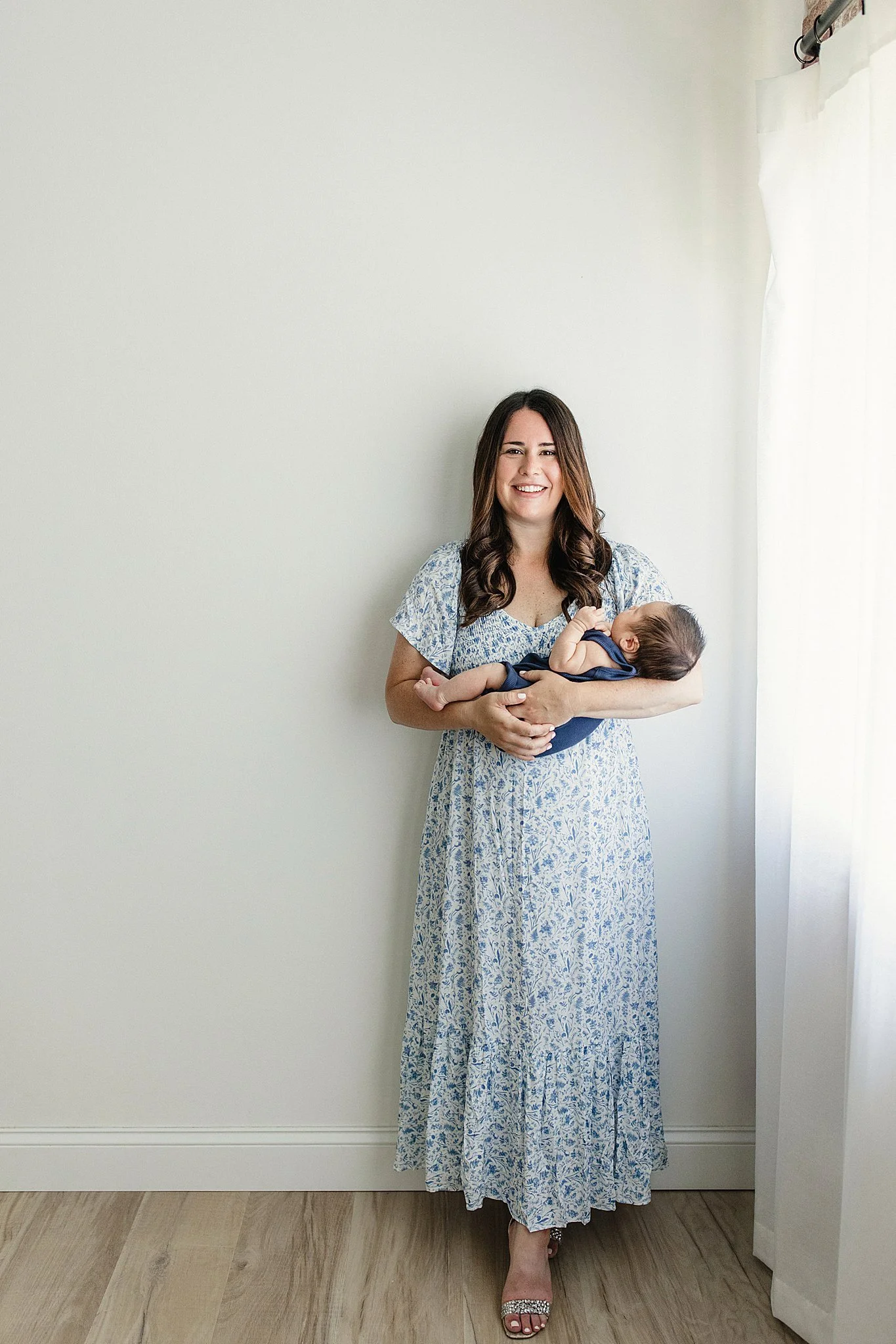 A woman wearing a long blue and white floral dress, standing barefoot on a wooden floor, holding a baby in her arms, with a white wall and sheer white curtains in the background during lifestyle newborn photos Naperville.