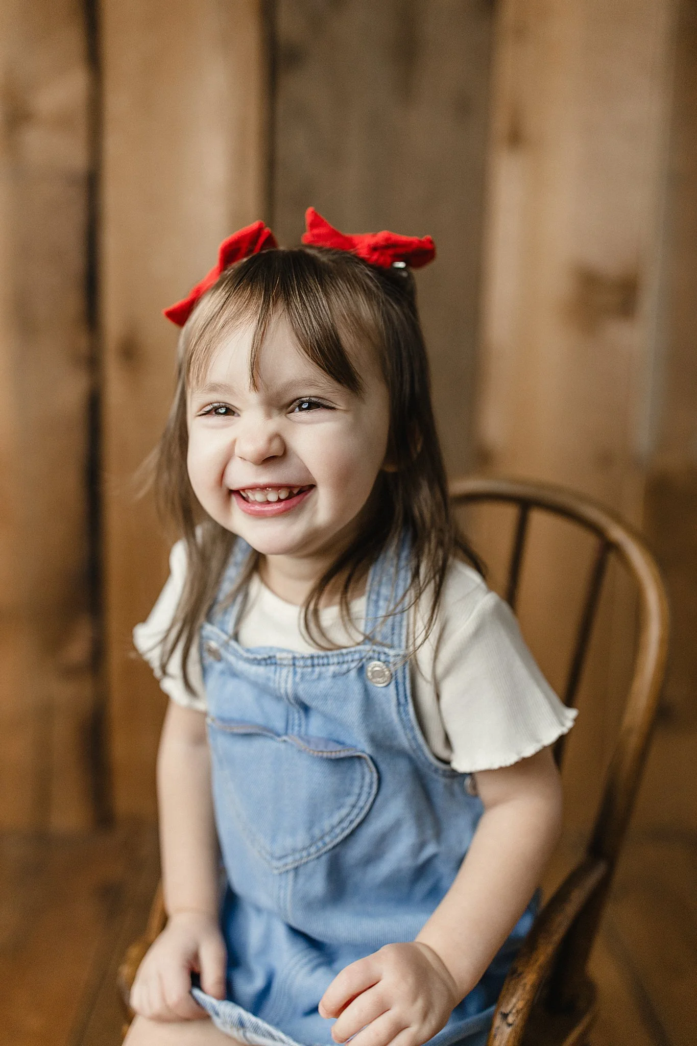 A young girl with brown hair, wearing a red bow, a white t-shirt, and denim overalls, sitting on a wooden chair and smiling for kids photos near me.