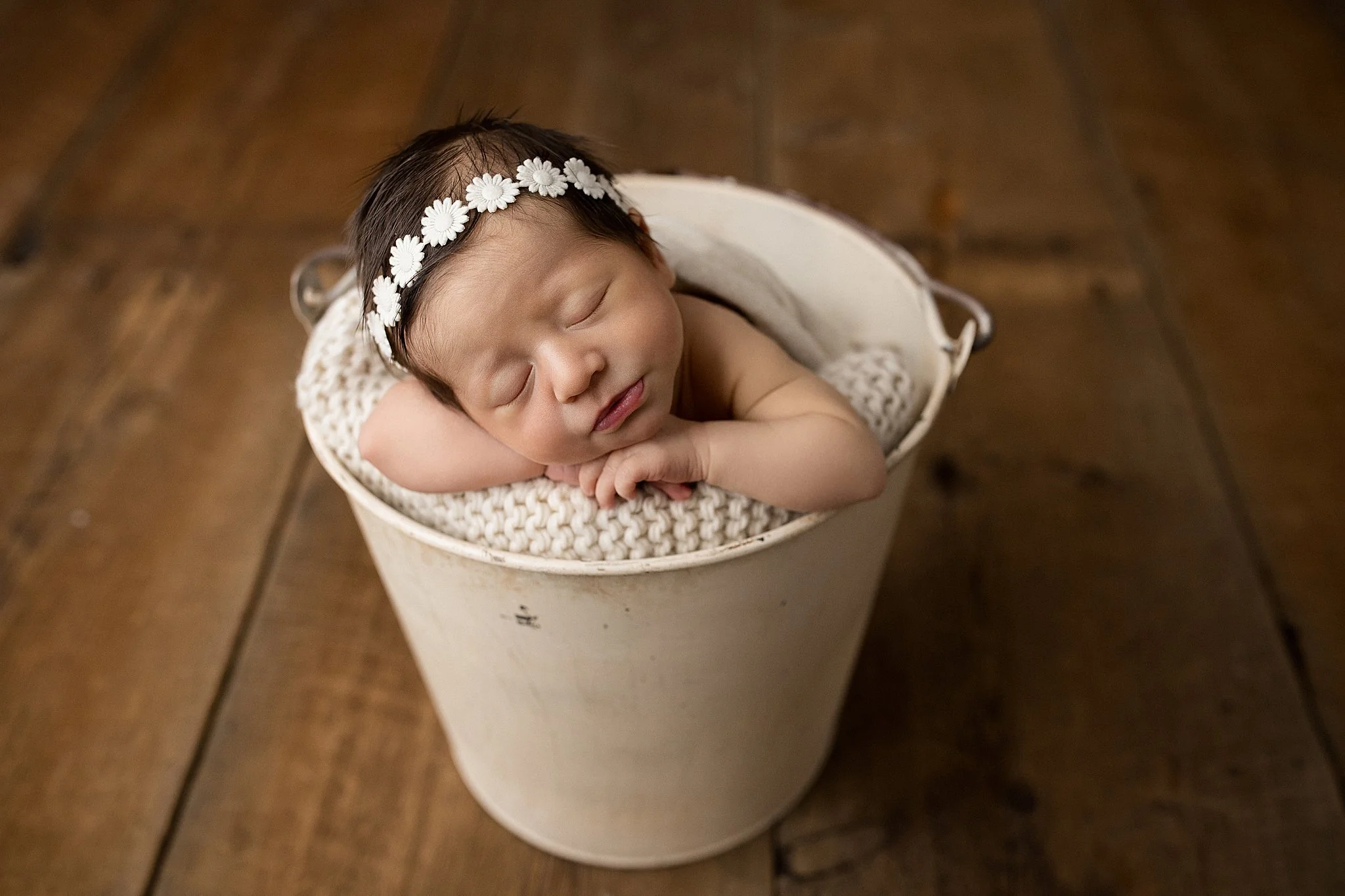 Young newborn baby with a flower headband sleeping inside a white bucket on a wooden floor, resting on a knitted blanket during a newborn session with Ally and B Photography.