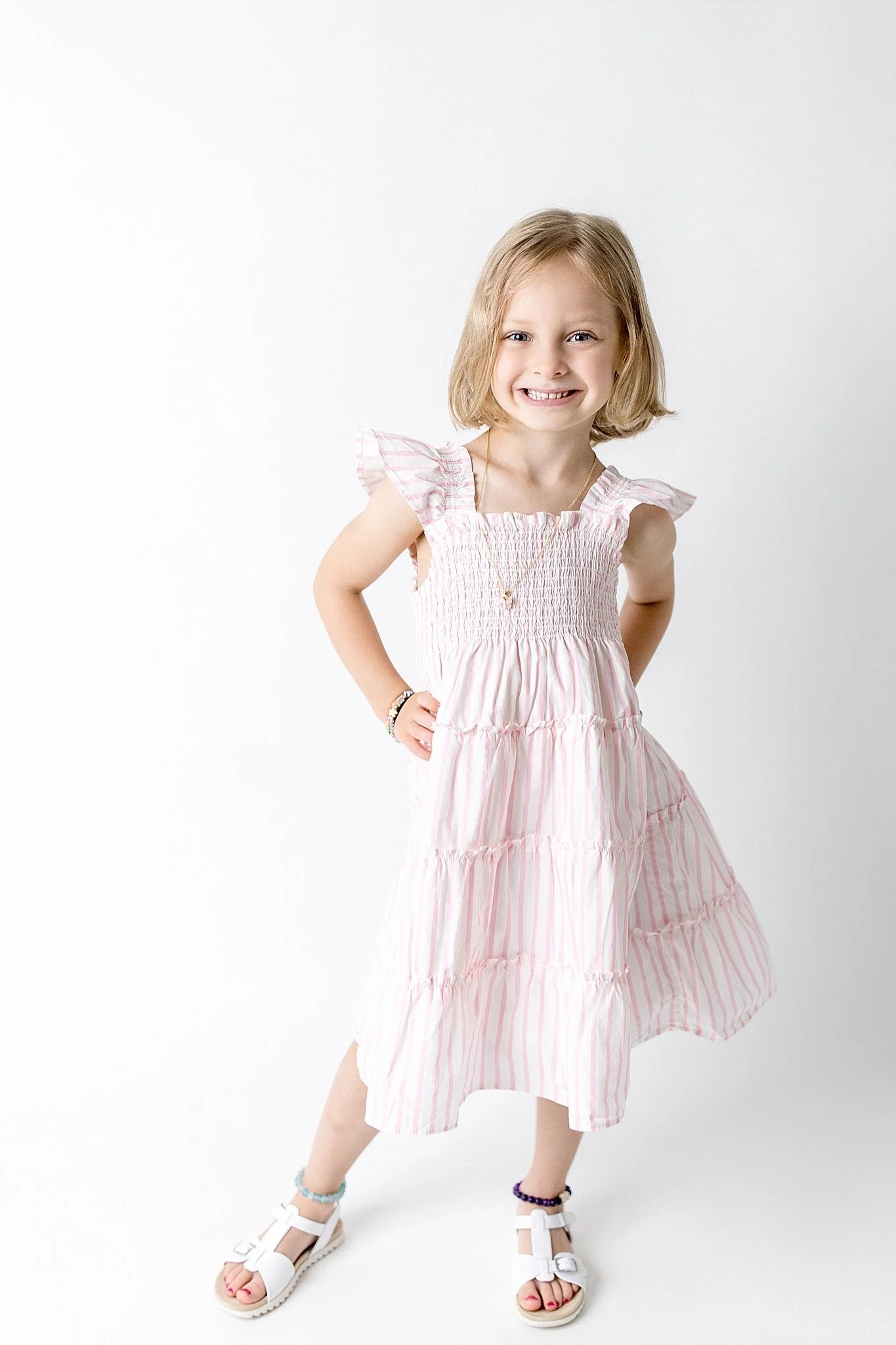 A young girl with blonde hair wearing a pink and white striped dress, sandals, and colorful bracelets, standing against a white background, smiling, with her hands on her hips during a personality mini session near me.