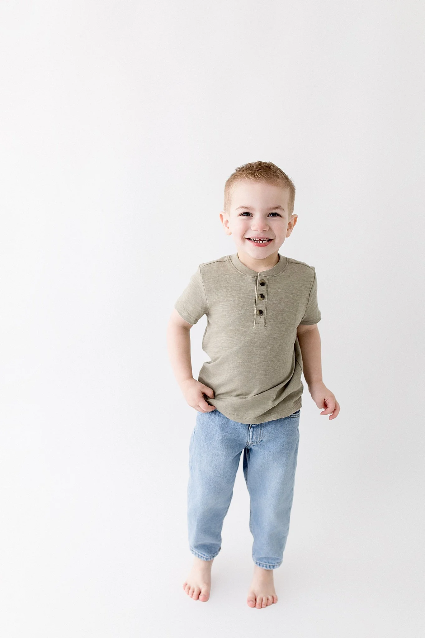 A young boy with short light brown hair, wearing a beige short-sleeve shirt and light blue jeans, smiling and standing barefoot against a plain white background with a childrens photographer near me.