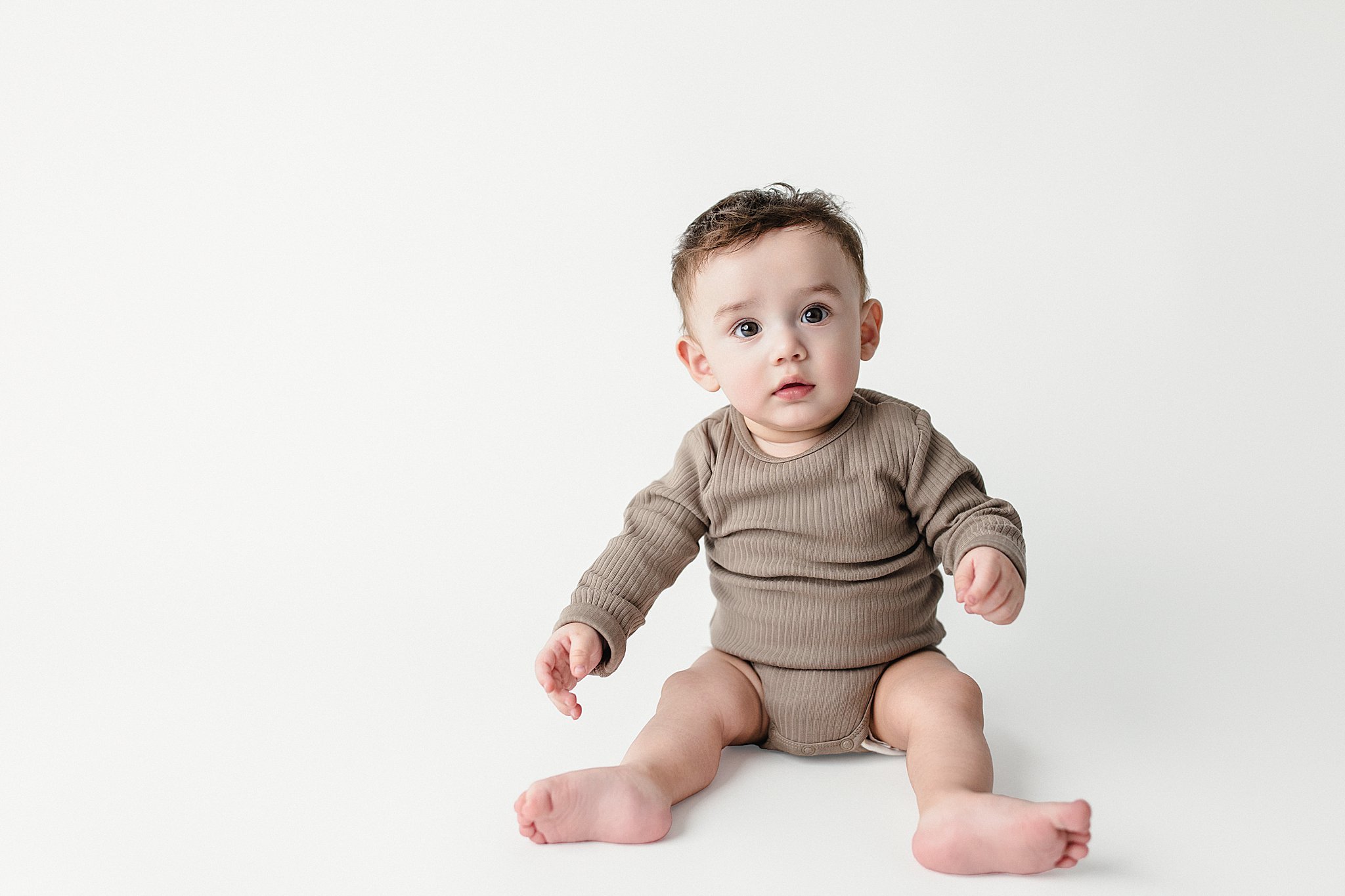 A baby with brown hair and big eyes sitting on a plain white background, wearing a brown long-sleeve shirt and matching diaper cover during baby photos with Ally and B Photography.