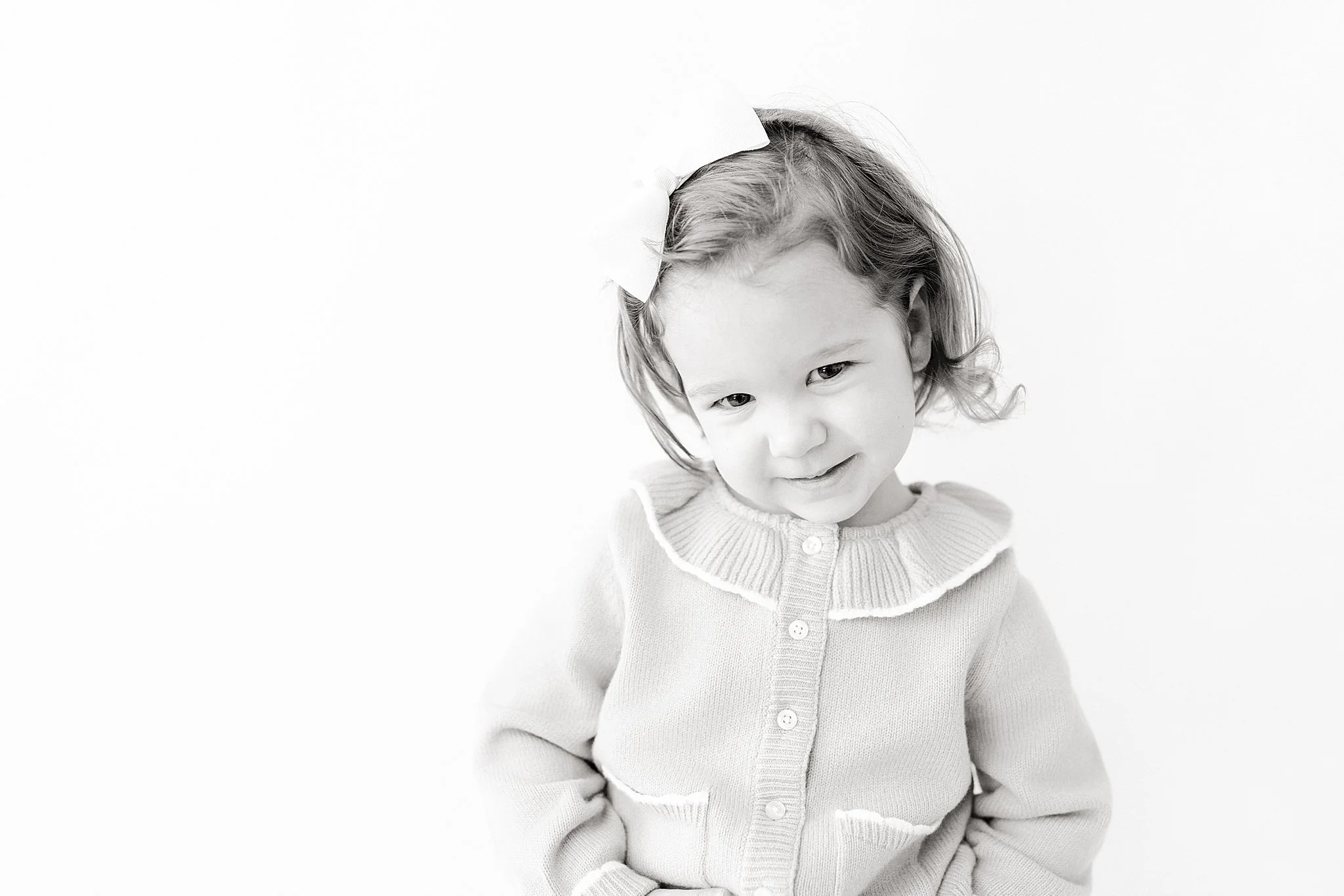 A young girl with a bow in her hair, wearing a sweater with a ruffled collar, smiling softly against a plain background for kids photos near me.