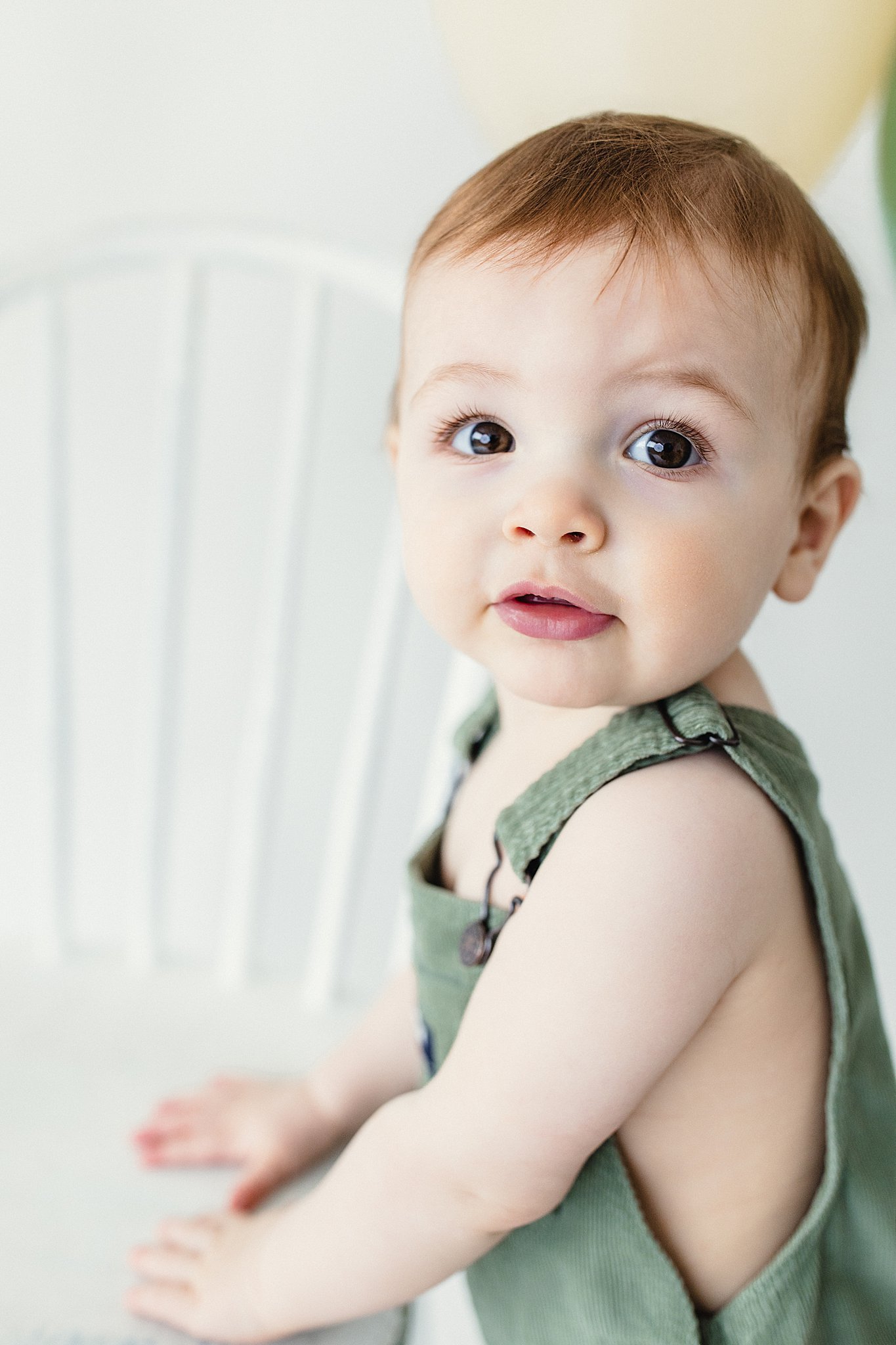 A young child with brown hair and big brown eyes, wearing a green sleeveless top, sitting on a white chair with a white background with best baby photographers in Naperville, IL.