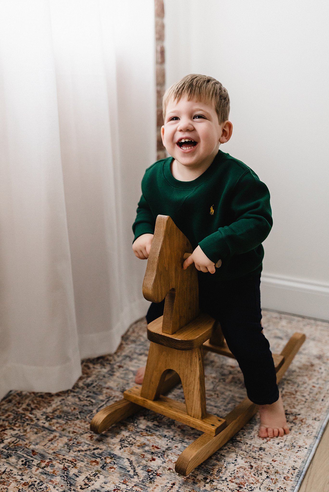 A young boy laughing while sitting on a wooden rocking horse during a mini milestone session in the Ally and B Photography studio in downtown Naperville, IL.