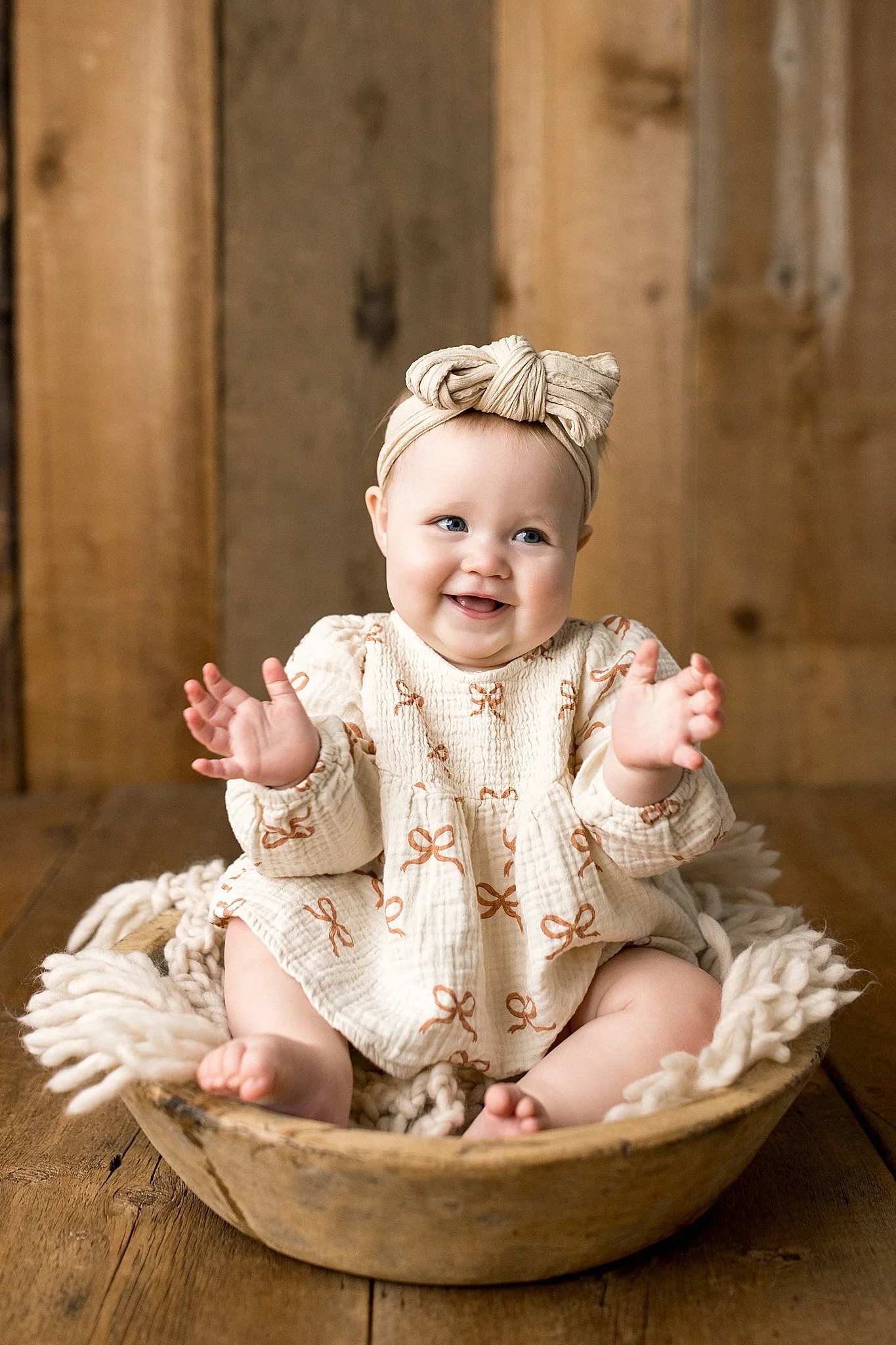 A baby girl sitting inside a wooden bowl on a wooden floor, wearing a beige dress with bow patterns and a headband with a bow, smiling and clapping her hands with the best baby photographer Naperville.