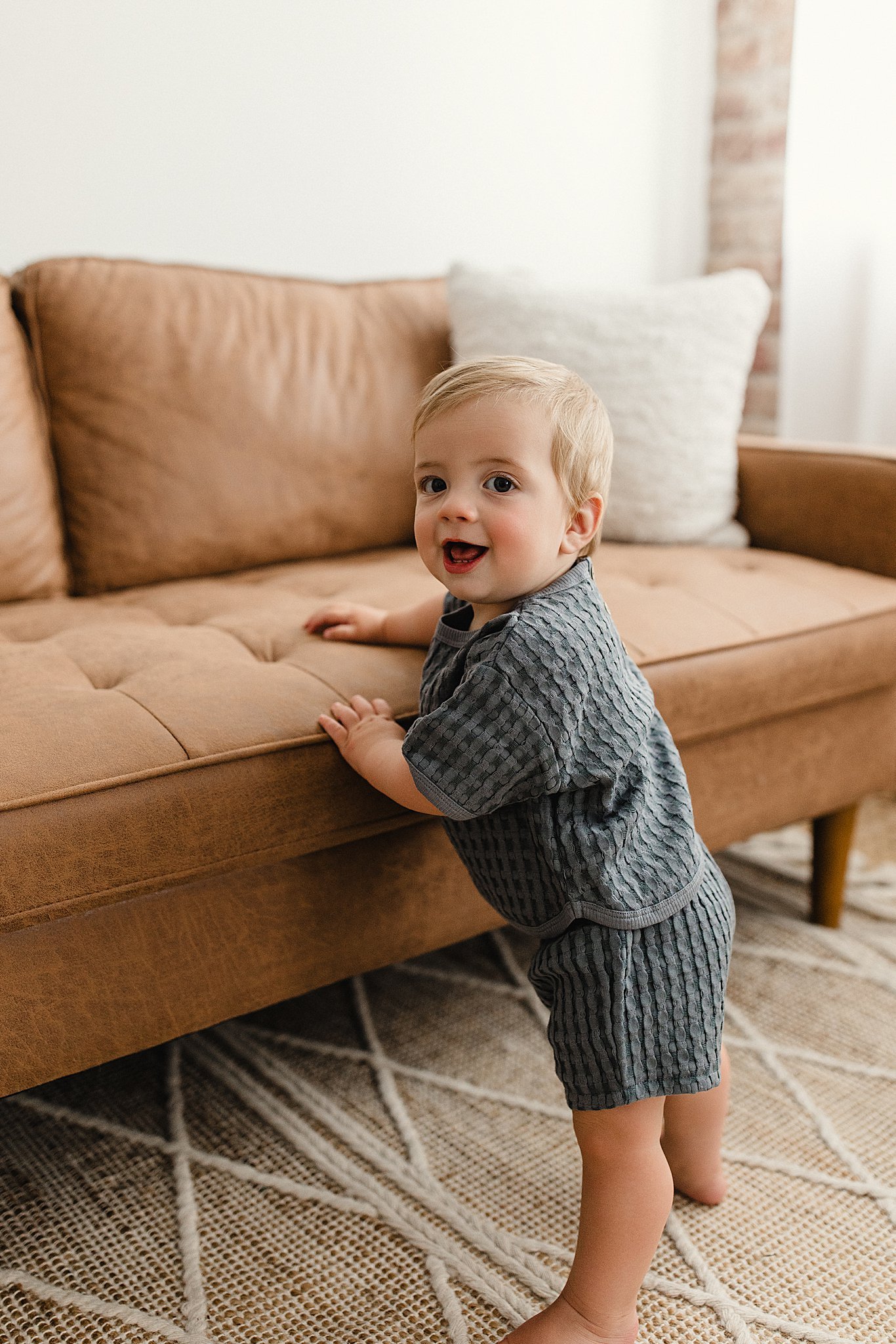 A young blonde-haired boy in a gray, textured outfit standing on a rug, holding onto the edge of a tan sofa with one hand and looking back at the camera with a smile for lifestyle photography near me.