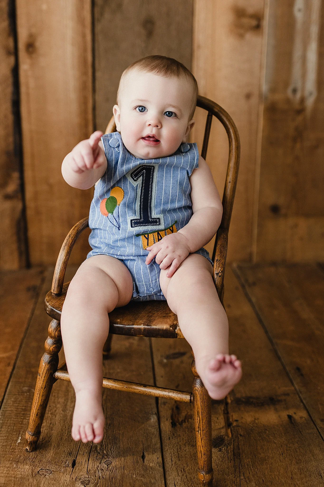 A baby sitting on a wooden chair, wearing a blue striped outfit with a large number one and colorful balloons on it, celebrating a first birthday during baby photos with the best baby photographer in Naperville, IL.
