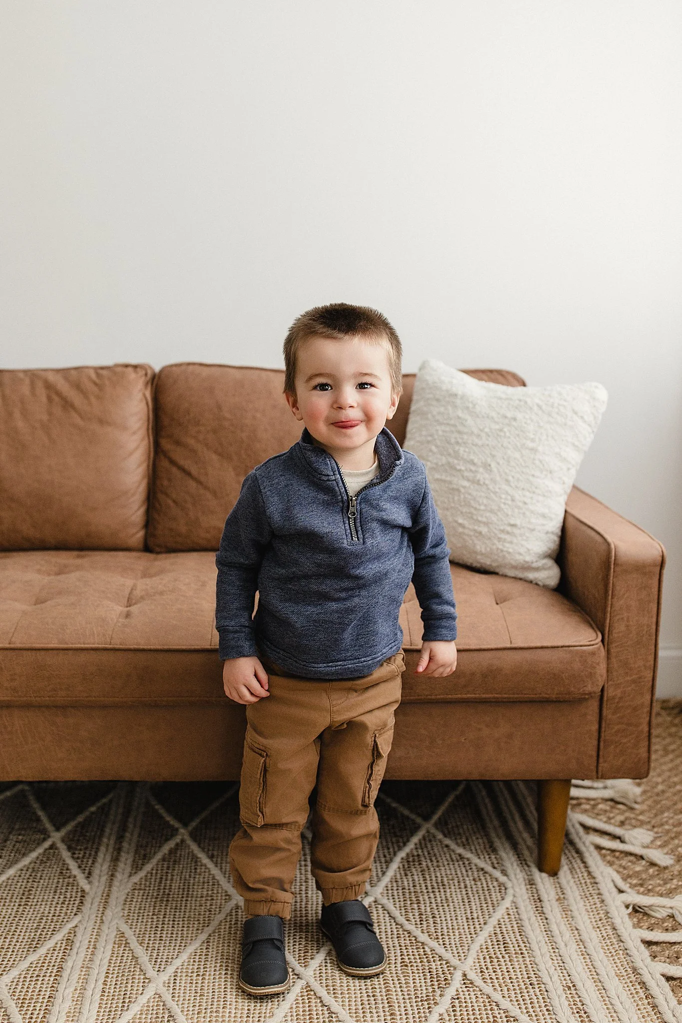 A young boy with short brown hair, wearing a blue zip-up sweater, tan cargo pants, and black shoes, standing in front of a brown sofa with a white pillow, on a patterned carpet in a living room during a kids photoshoot in Naperville.