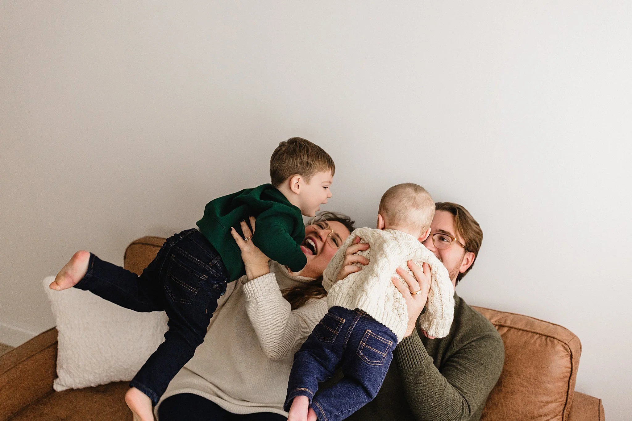 Family of four playing on a brown couch in living room, two children and two adults, all smiling and enjoying quality time together during a downtown Naperville studio family photoshoot.