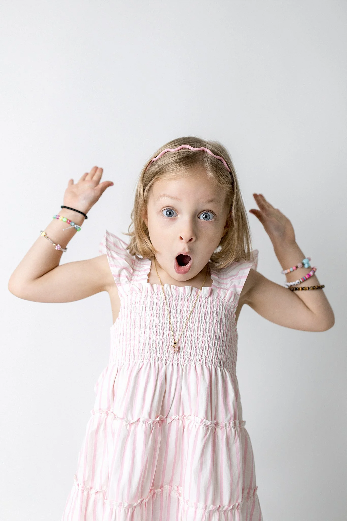 A young girl with blonde hair, blue eyes, and an expression of surprise or excitement, wearing a pink and white striped dress and a pink headband, with her hands raised near her head against a plain white background for personality sessions near me.