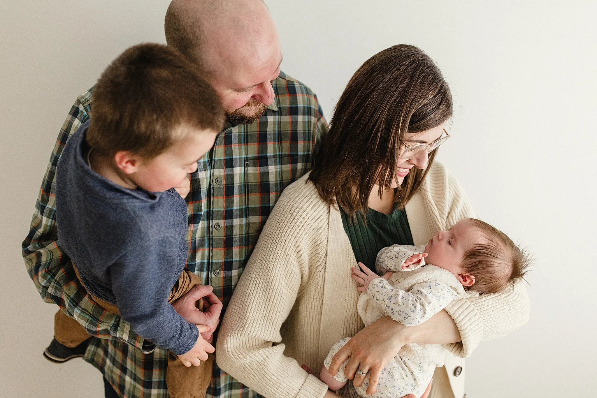 A woman holding a baby, surrounded by a man and a young boy, all smiling and looking at the baby, in a family portrait with family photographers near me.