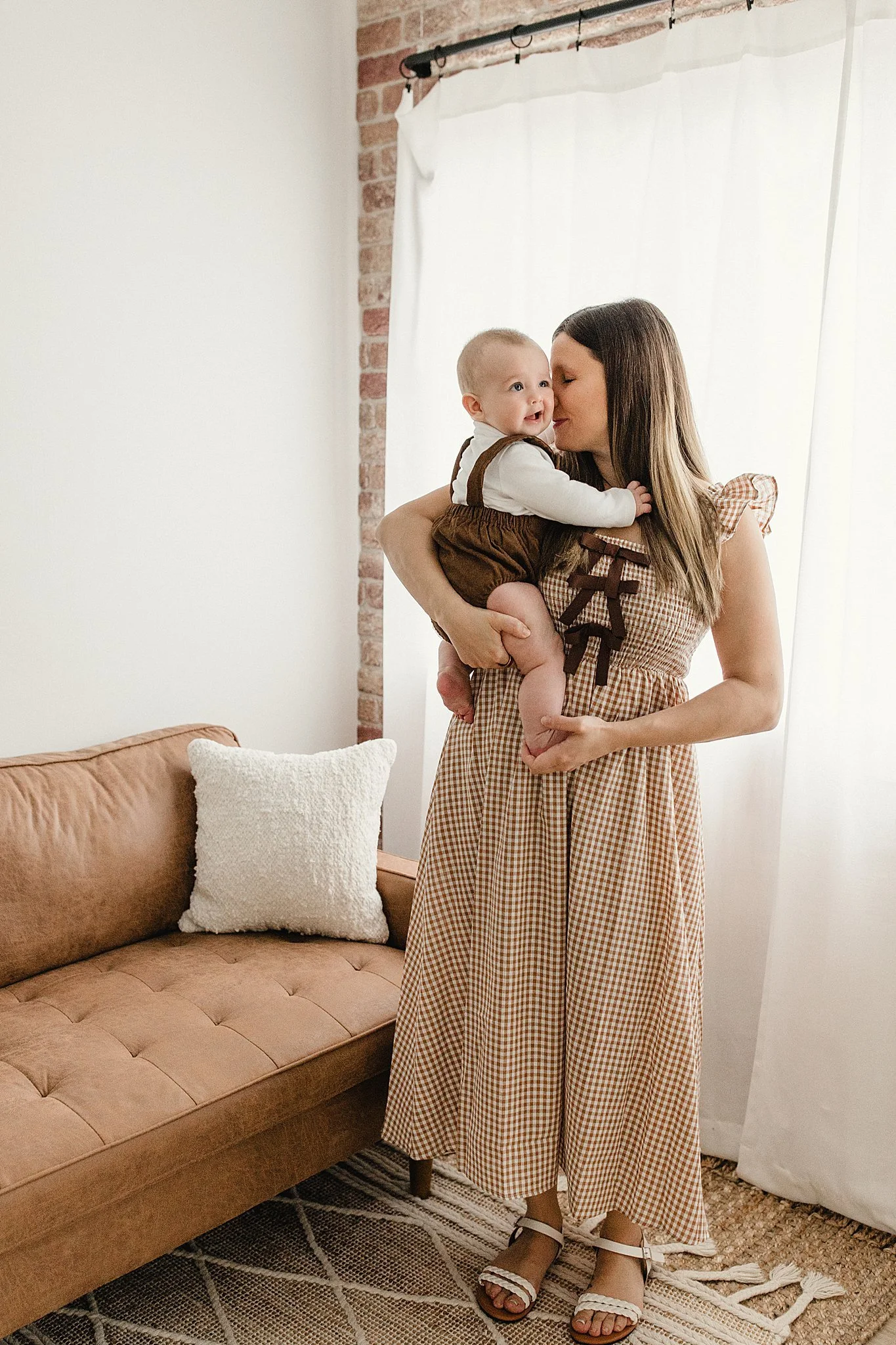 A woman holding a smiling baby in her arms inside a room with a brick wall and white curtains for family lifestyle photos with Ally and B Photography.