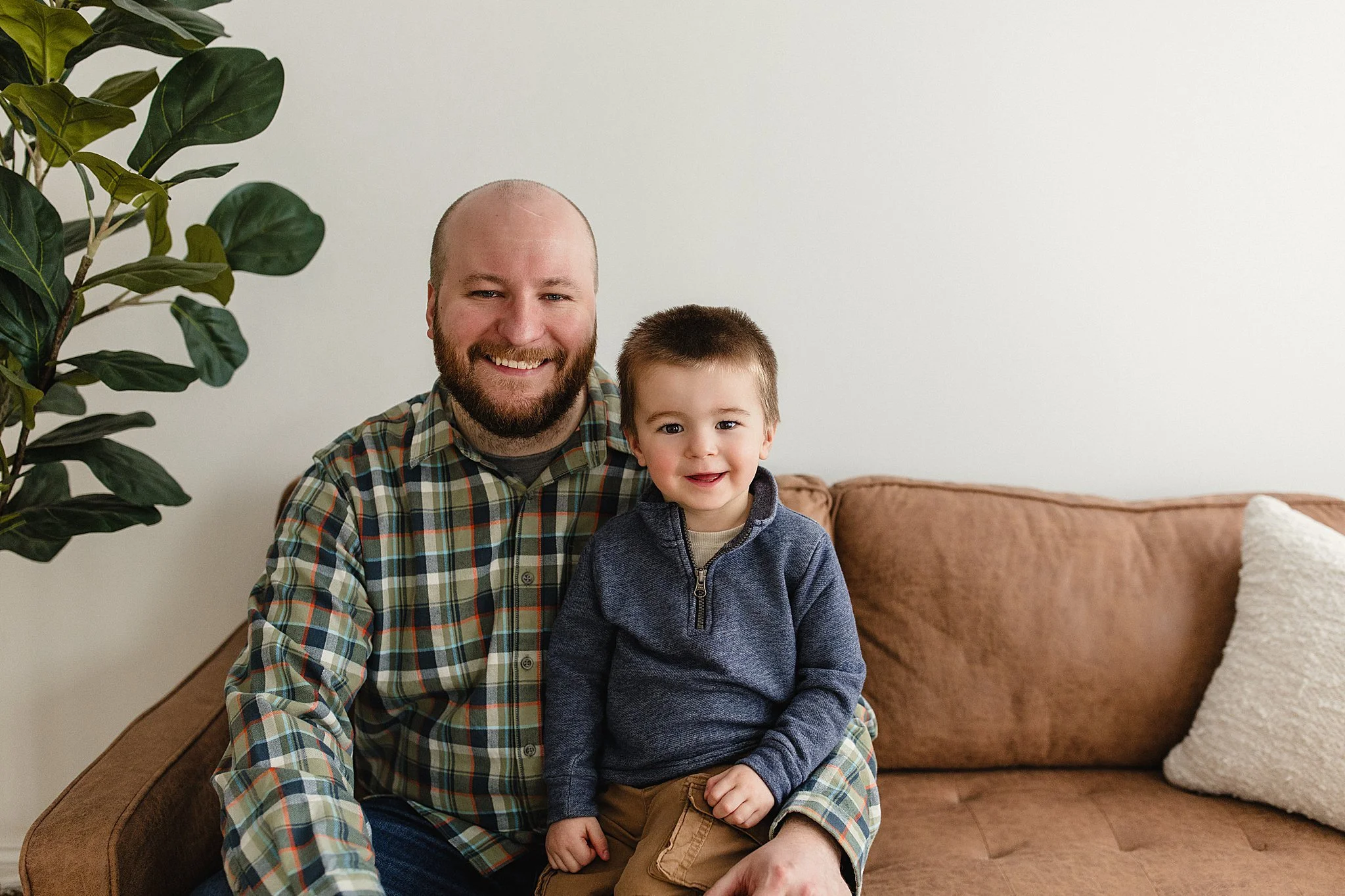A smiling man and a young boy sitting together on a tan sofa, with a large green plant on the left side of the image and a white pillow on the right, in a cozy living room in a photo studio near me.