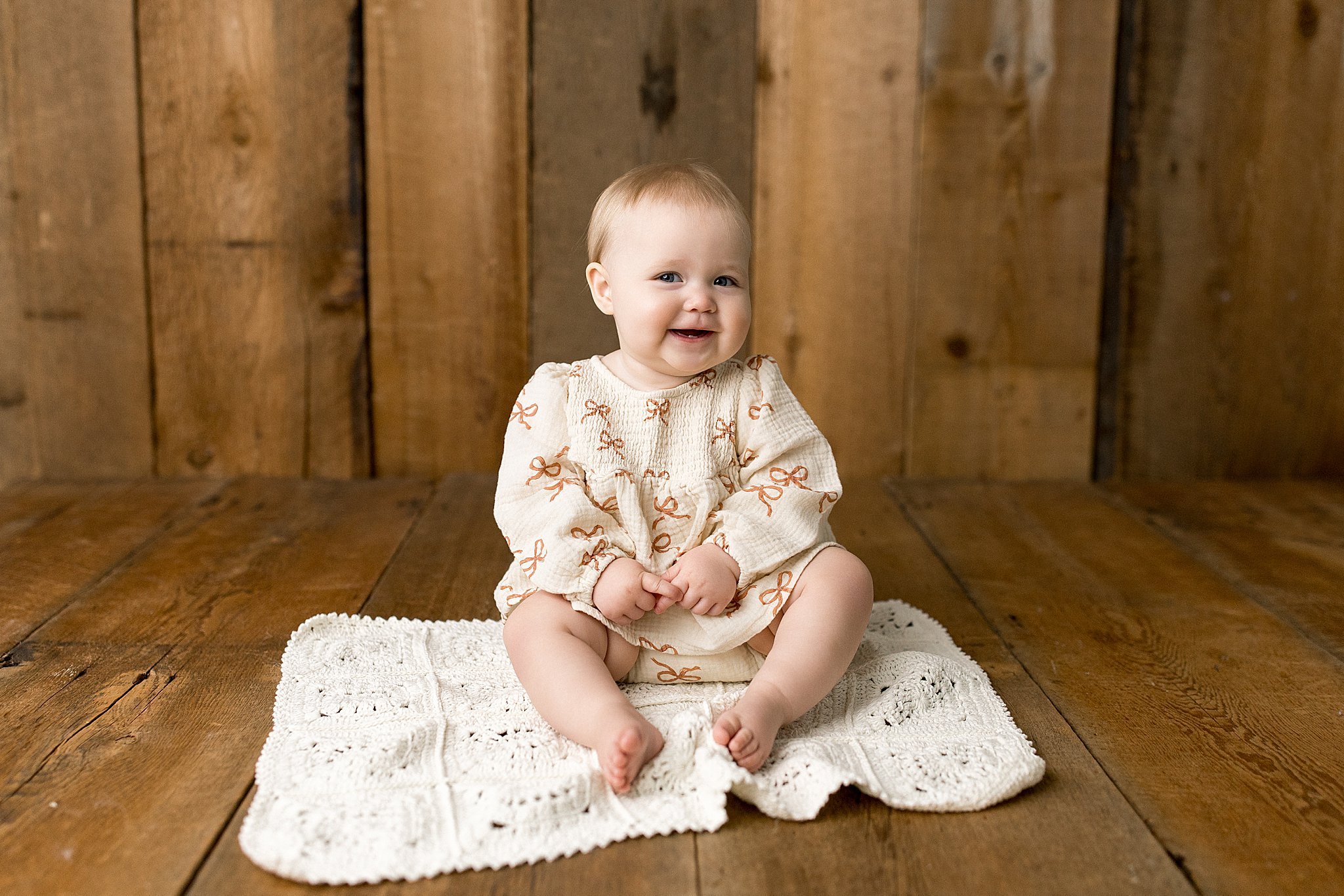 A smiling baby girl sitting on a white crocheted blanket on a wooden floor with a wooden wall background with Marie Lopez photographer in Naperville, IL.