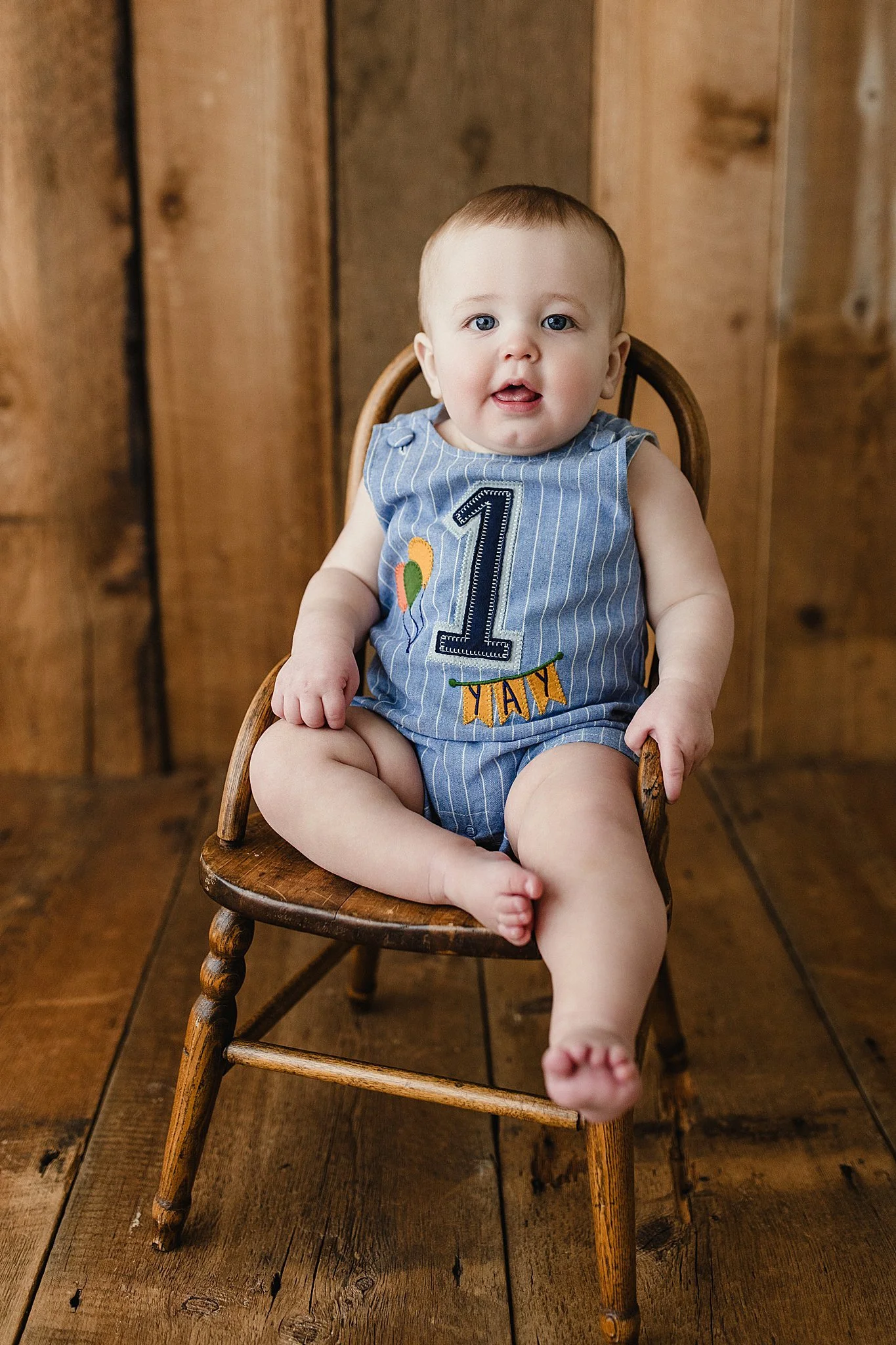 Baby sitting on a wooden chair wearing a blue outfit with a number 1 and the words '1st Birthday' on it with Marie Lopez photographer of Ally and B Photography in Naperville, IL.