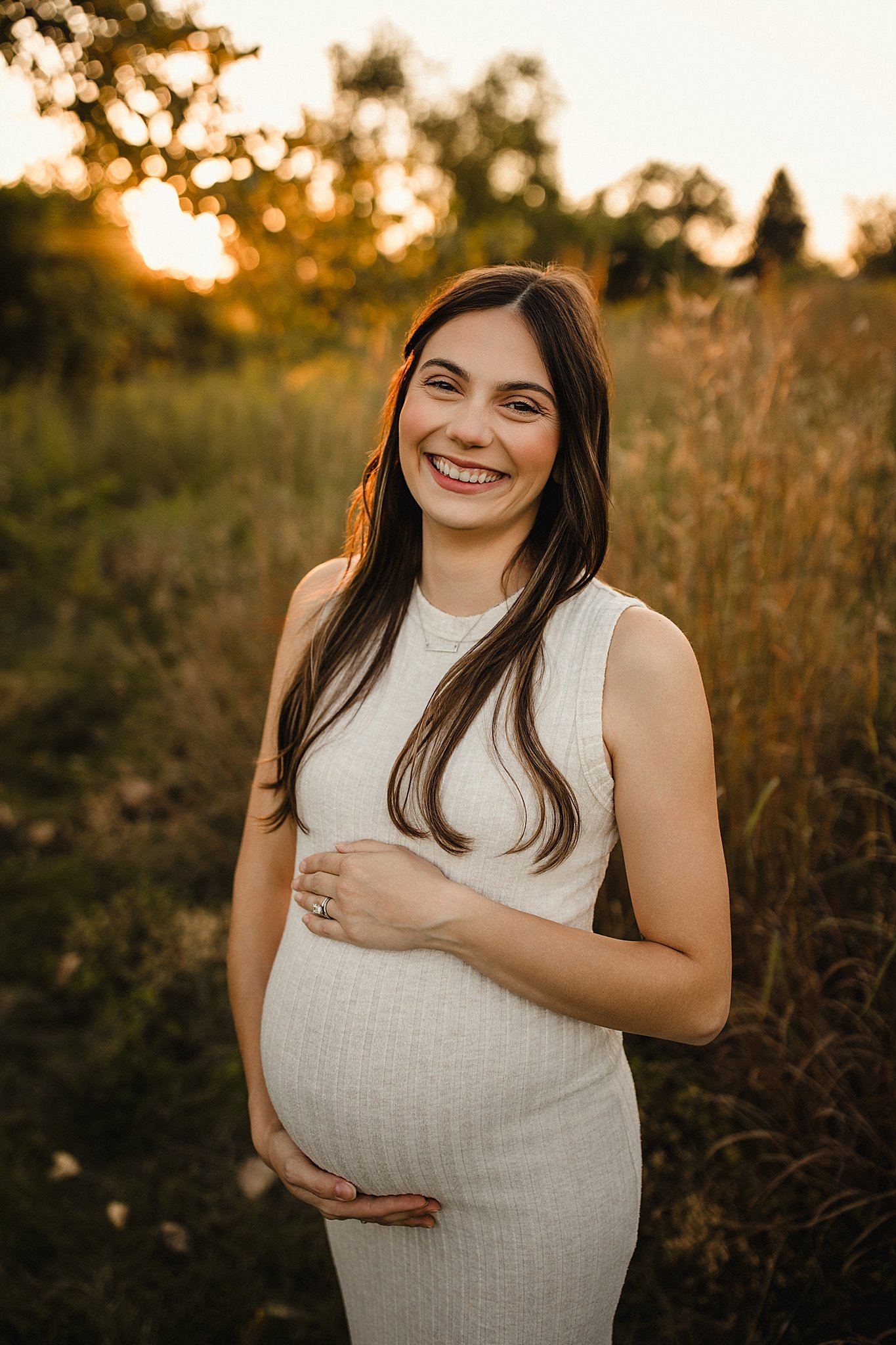 A smiling pregnant woman with long dark hair in a sleeveless beige dress standing outdoors in a field at sunset, with trees and a golden sky in the background during a photo session with the best Naperville maternity photographer.