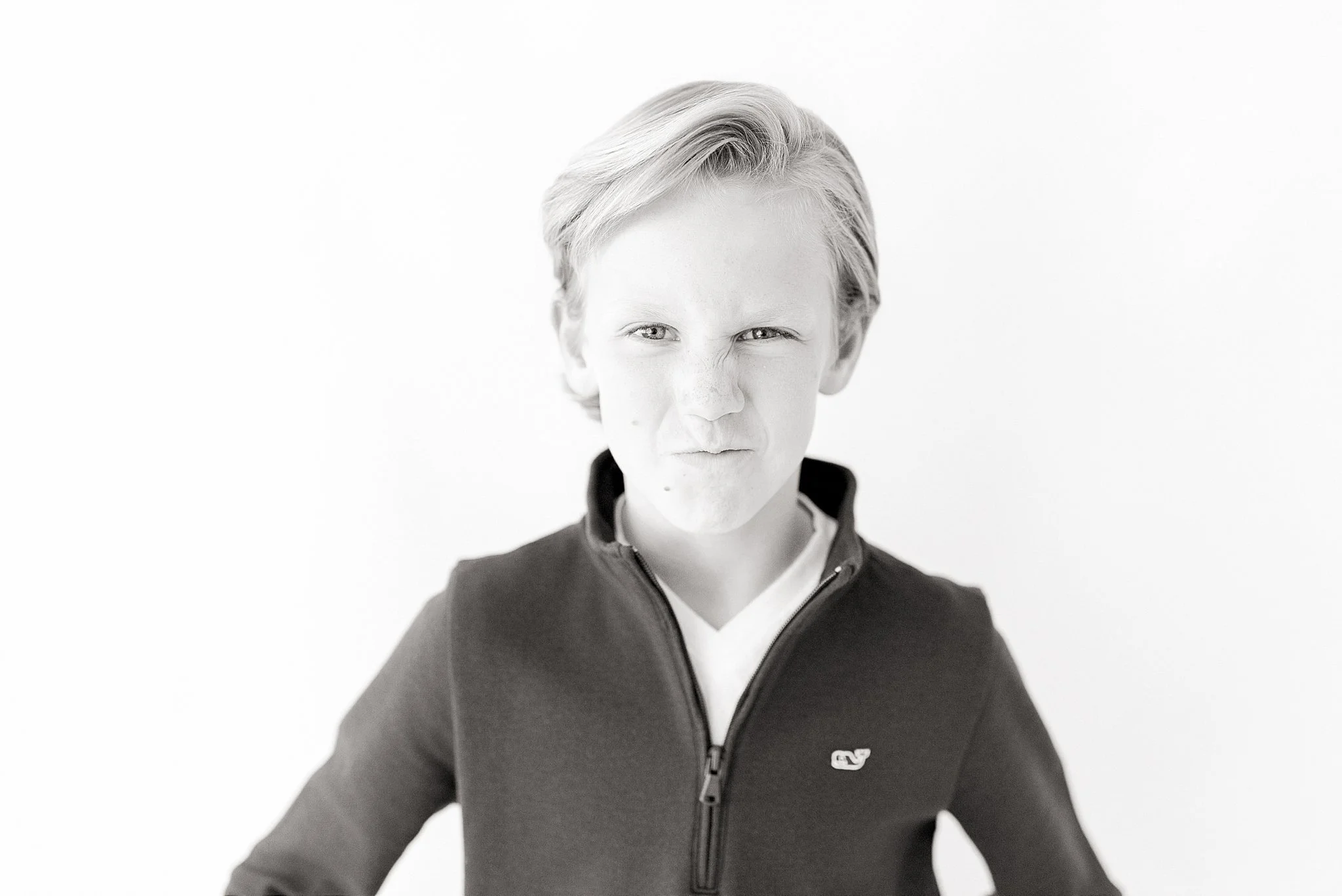 A young boy with blonde hair styled back, making a playful, scrunched face, wearing a zip-up dark jacket with a small logo on the chest, standing against a plain light background during Naperville personality minis.