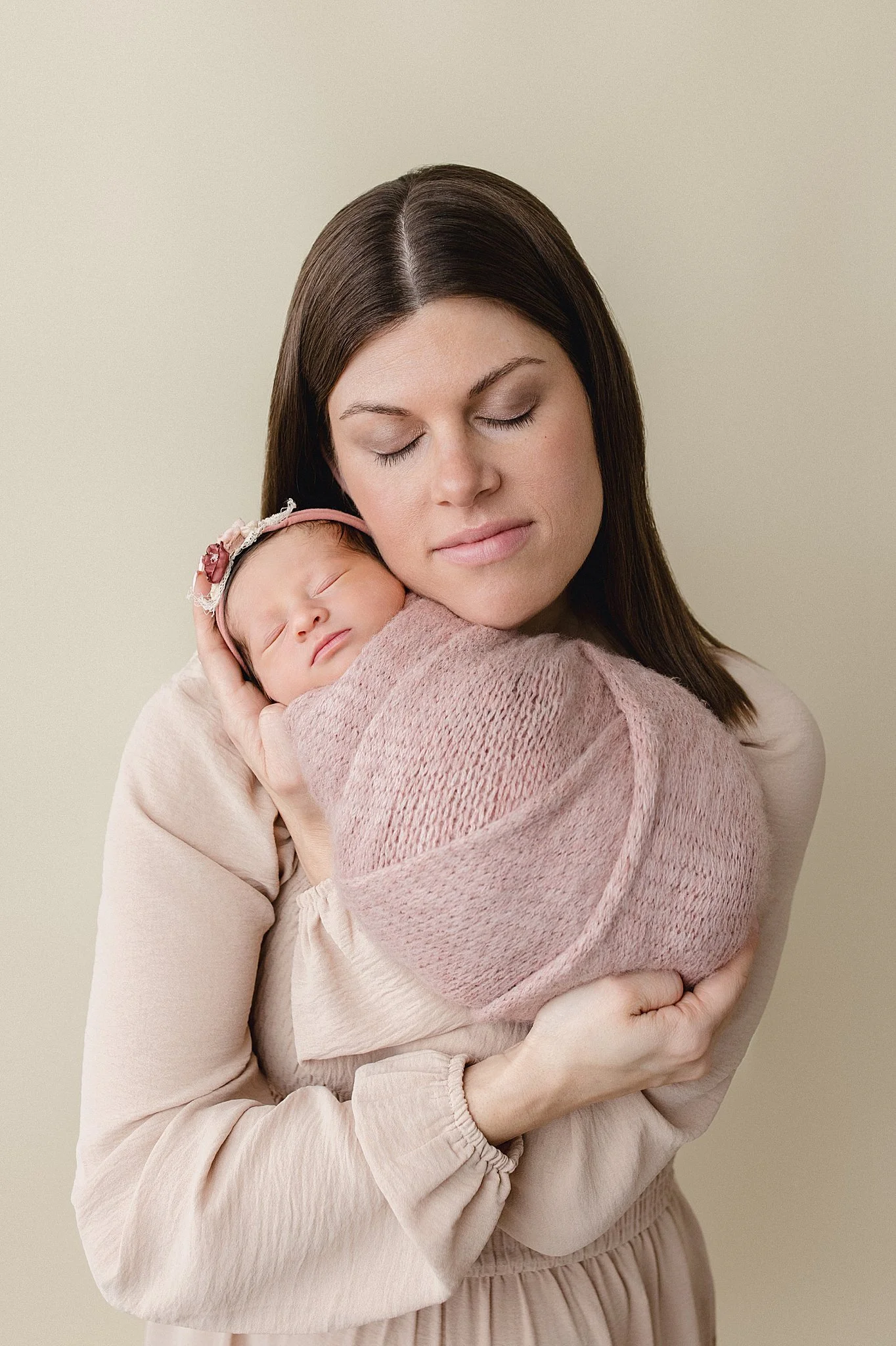 A mother with long brown hair gently holding and cuddling her sleeping baby wrapped in a pink blanket, against a neutral background.