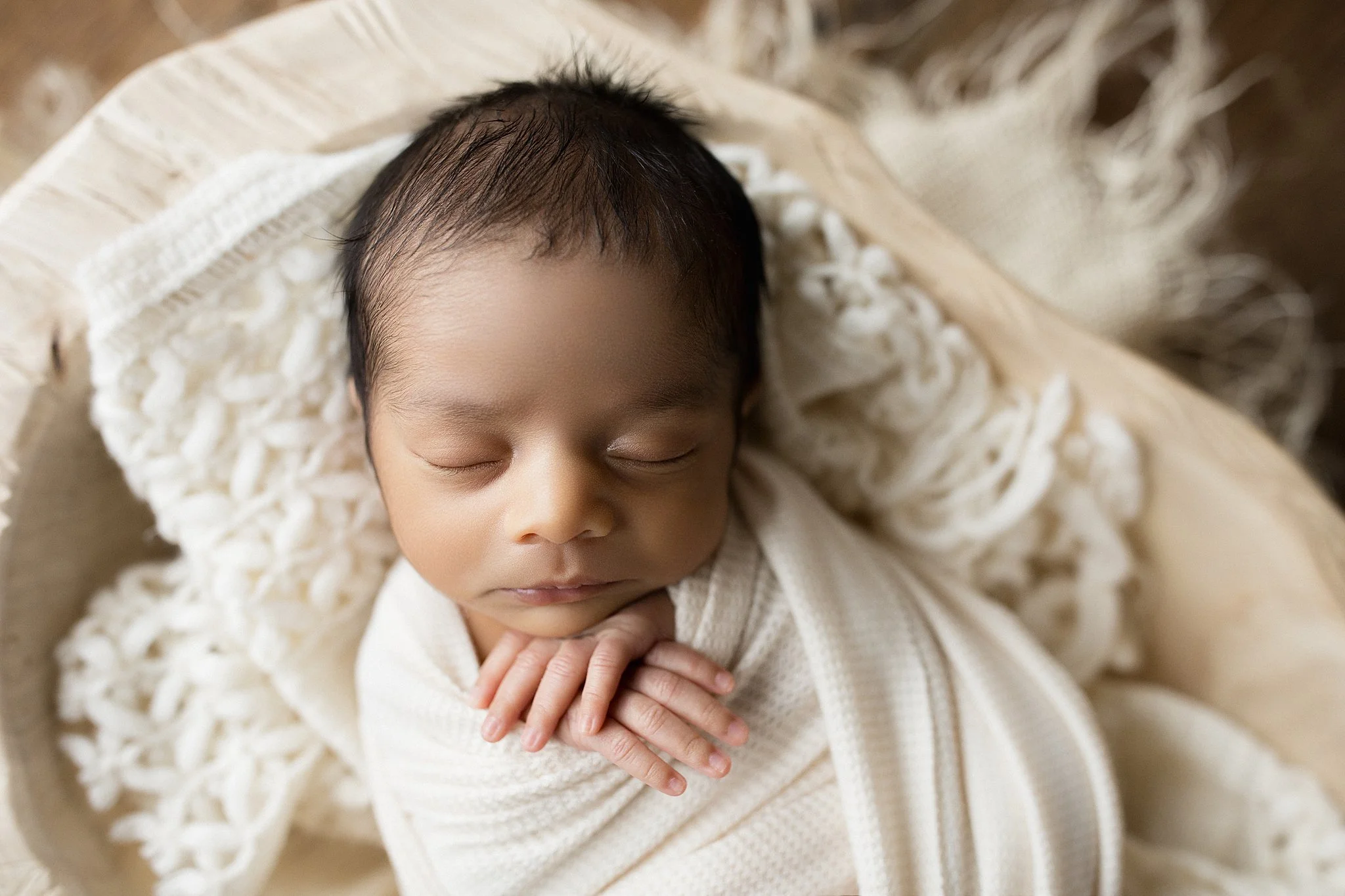 A sleeping newborn baby wrapped in a cream-colored blanket, lying on a textured cream and beige fabric surface during a posed newborn photography session.