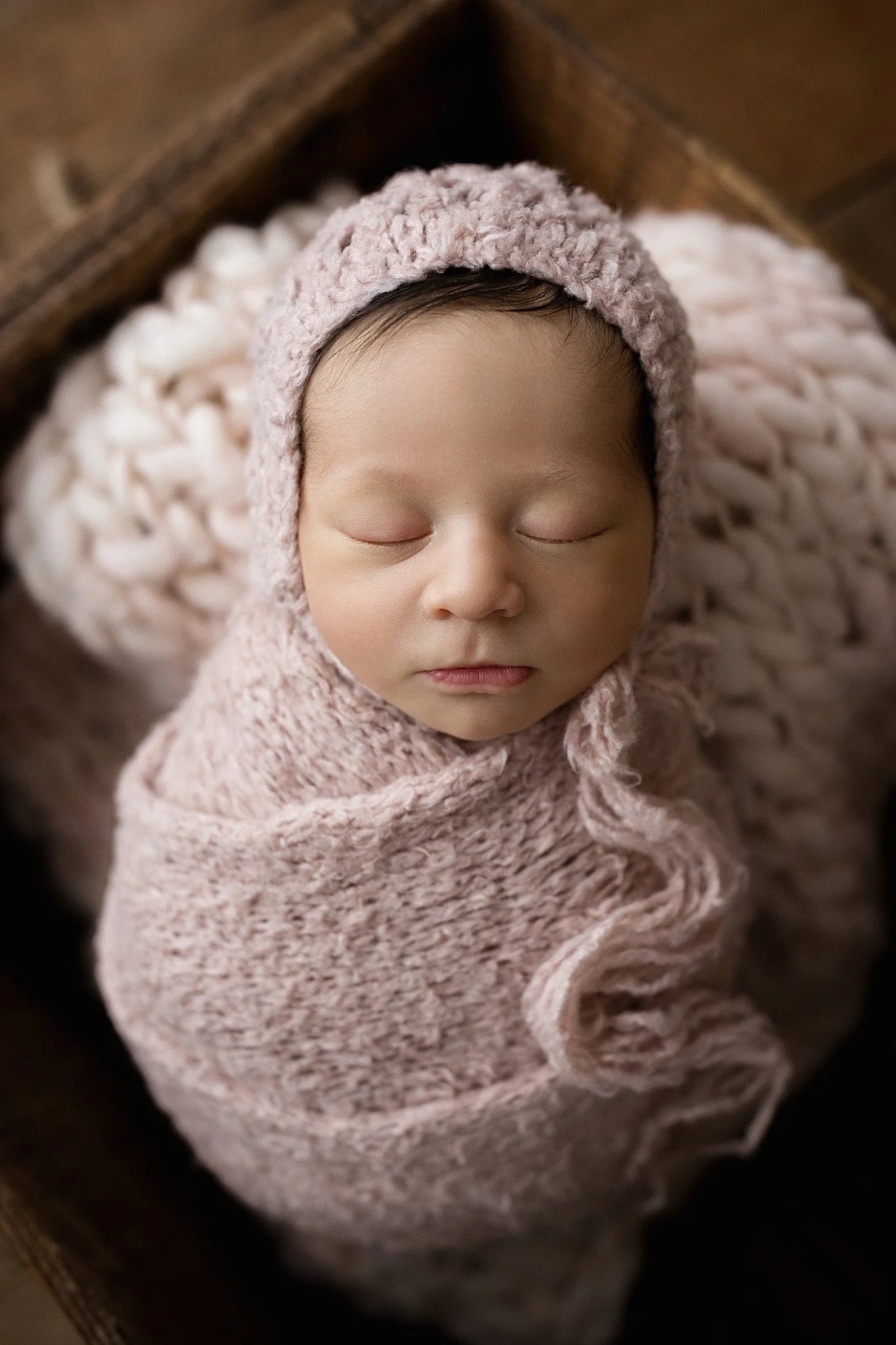 A sleeping newborn baby wrapped in a pink knitted blanket, wearing a matching pink knitted hat, lying on a soft pink blanket inside a wooden box with best newborn photographer Naperville, IL.