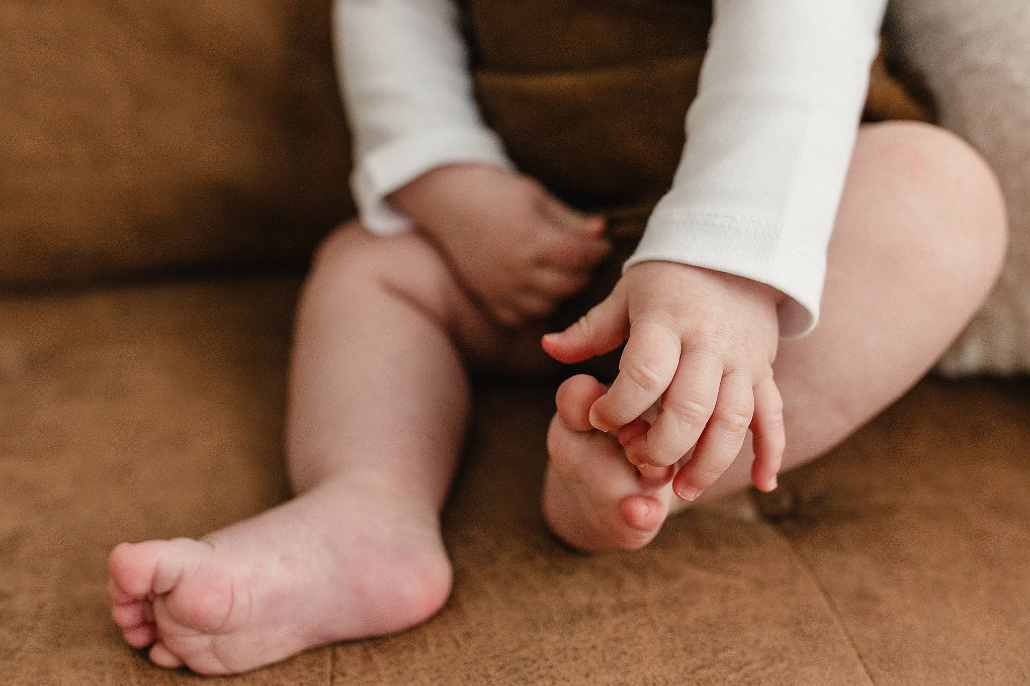 Close-up of a child's legs with hands holding their feet, sitting on a brown surface during baby photos near me.
