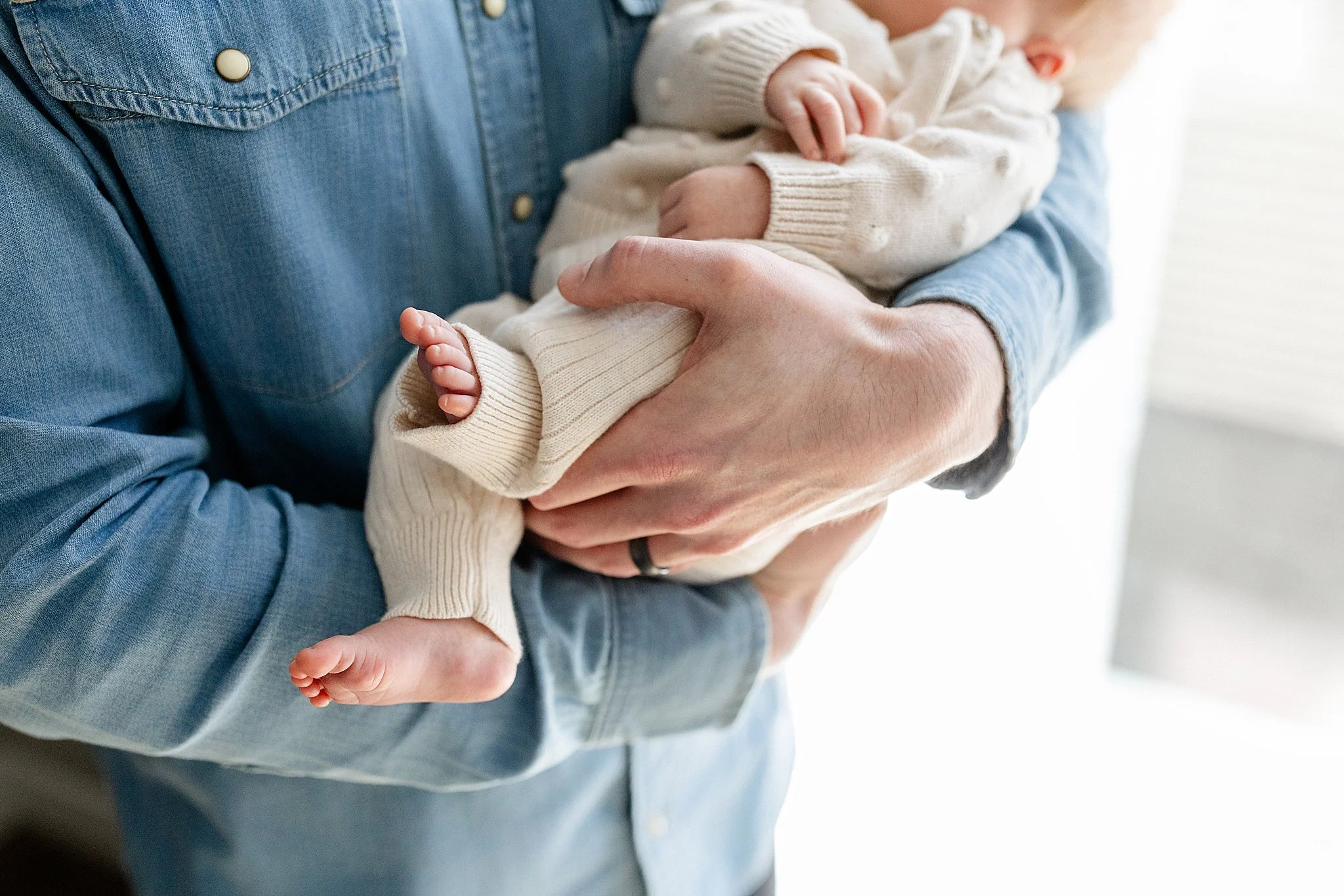 Person holding a baby dressed in cream-colored sweater and pants, with the focus on the hands and feet with Naperville lifestyle photographers.