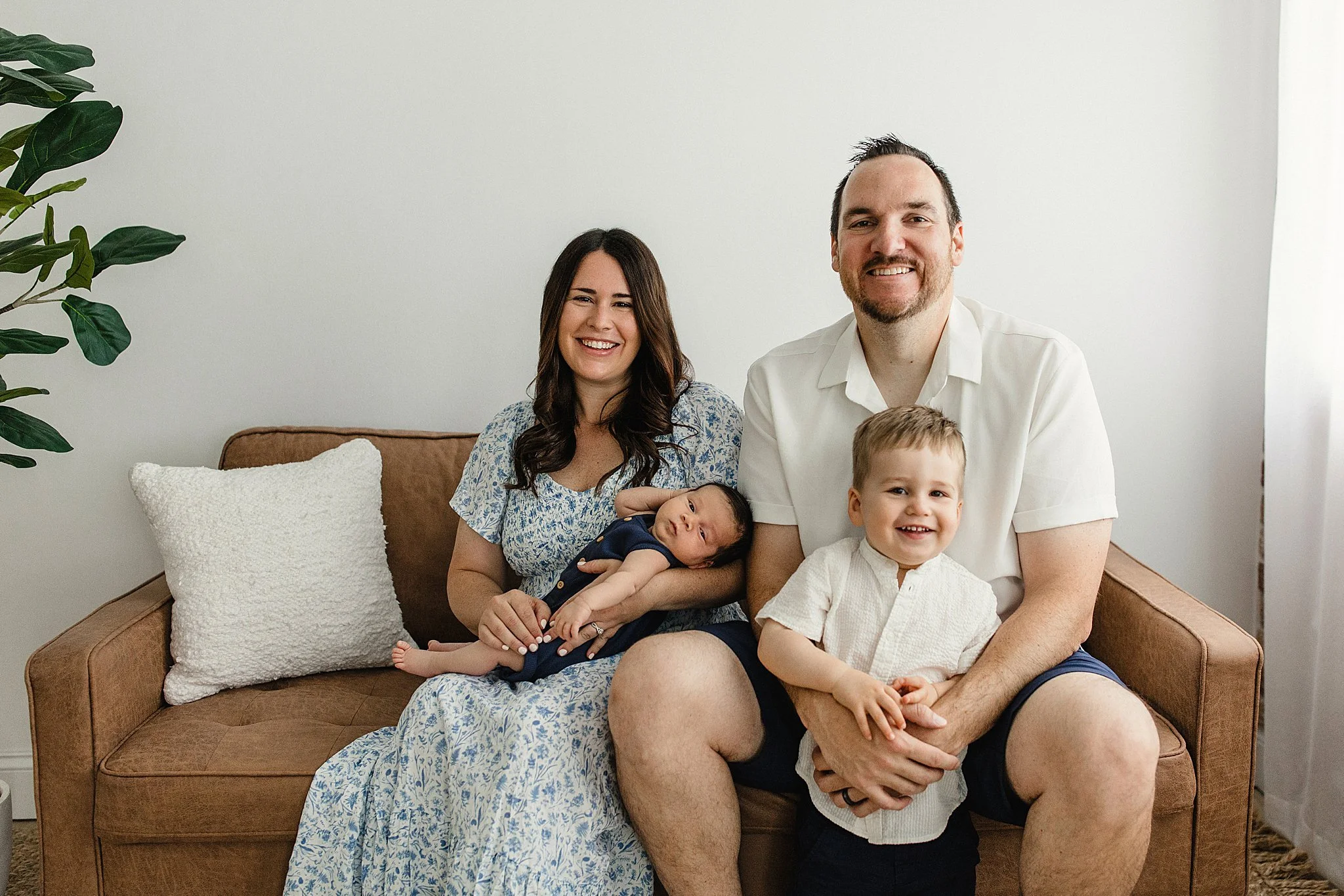 A smiling family of four sitting on a brown sofa in a bright room, with a woman holding a baby and a man sitting with a young boy during lifestyle newborn photography Naperville.