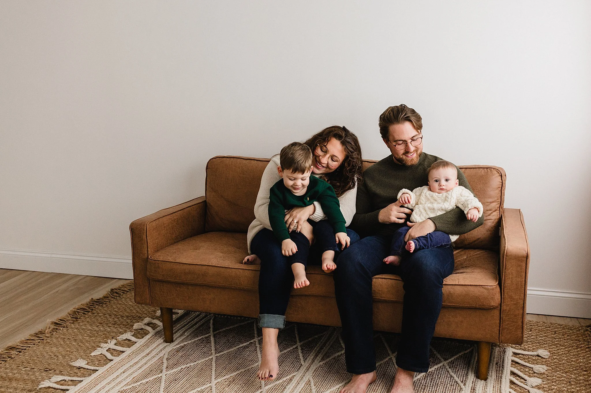 A happy family of four sitting on a brown couch in a cozy living room, playing and laughing together during a studio family photography session with Ally and B Photography in downtown Naperville, IL