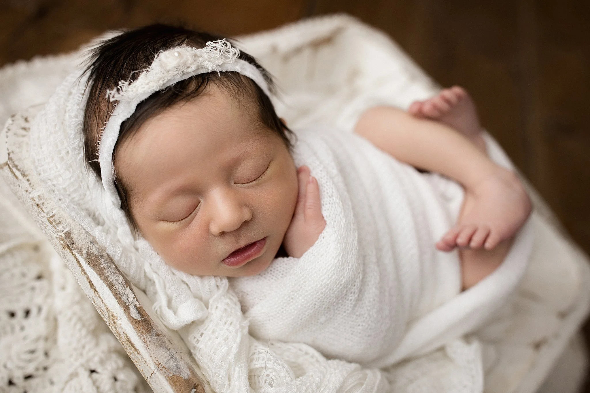 A sleeping newborn baby wrapped in a white blanket and wearing a white headband, resting on a white crocheted blanket.