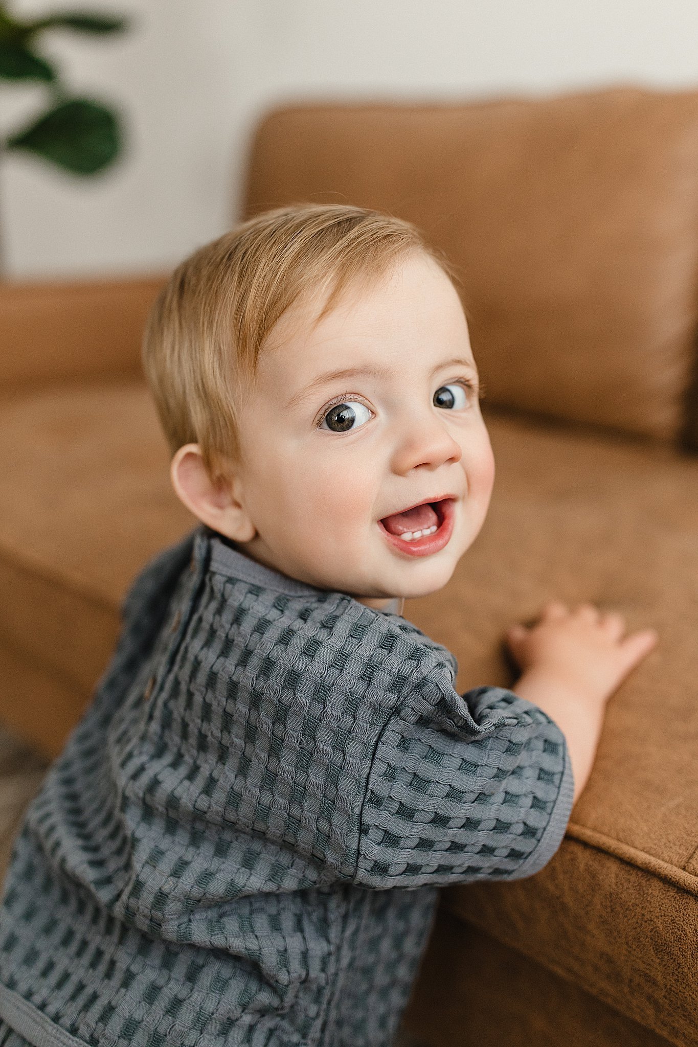 A young child with light hair and big blue eyes looking up with an open mouth, sitting on a brown couch for infant photography near me.