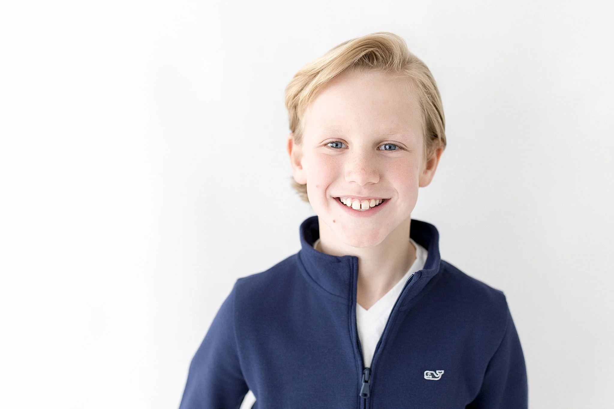 A young boy with blonde hair, blue eyes, and freckles, smiling while wearing a navy blue zip-up jacket against a white background with a children's photographer near me.