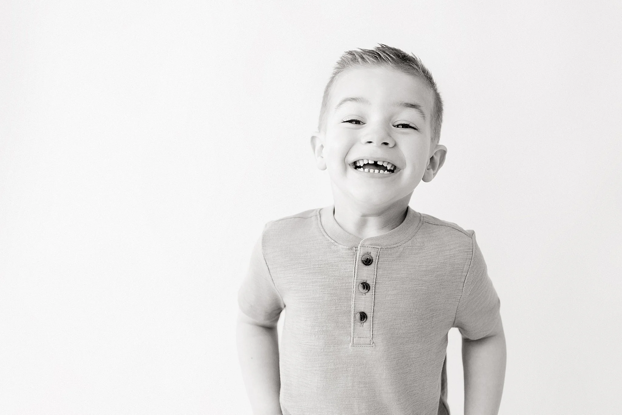 A young boy smiling with gap teeth, wearing a light-colored shirt with three buttons near the collar, and standing against a plain light background with kids photographers near me.