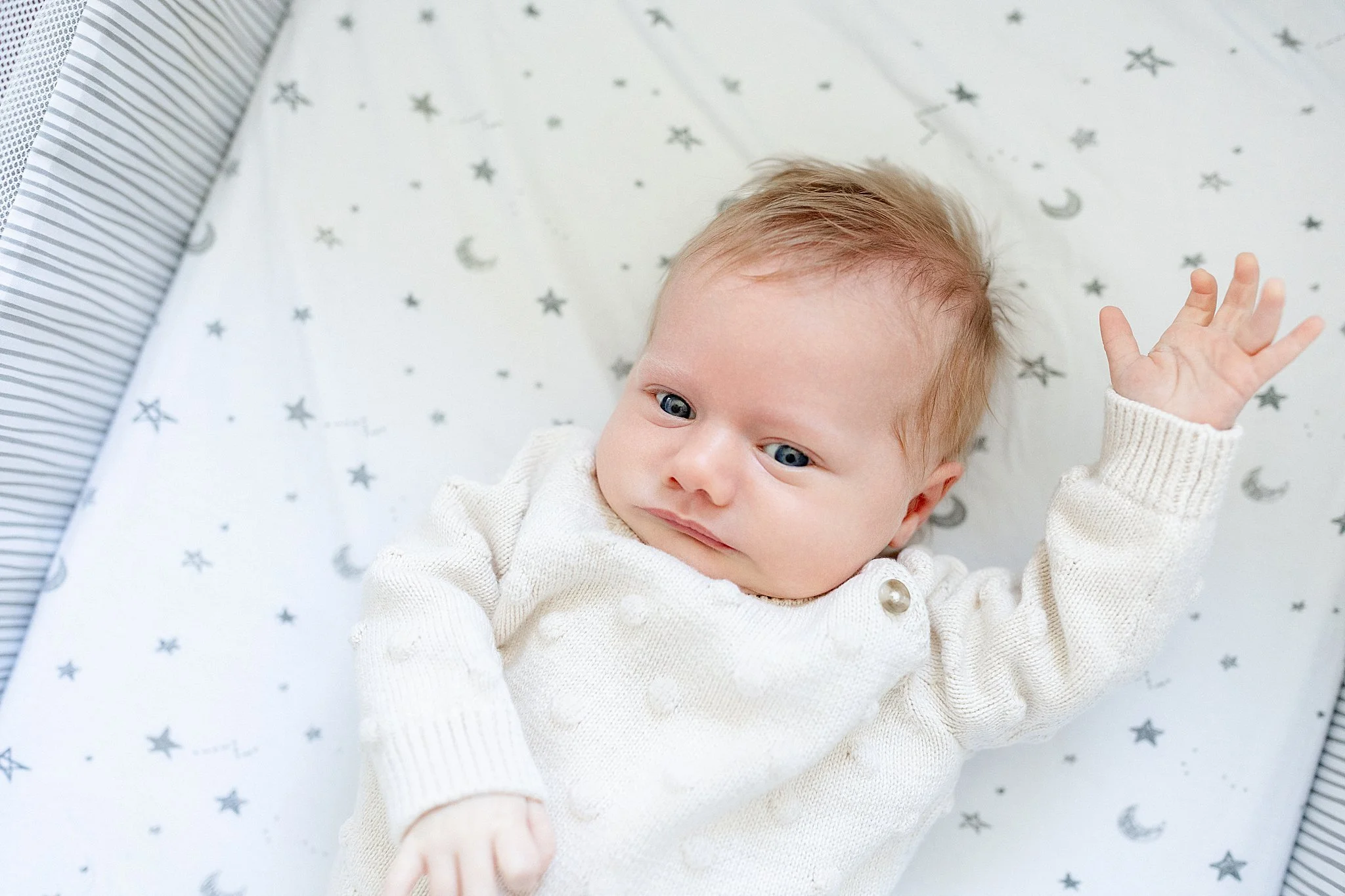 A baby with blue eyes and light hair lying on a star-patterned sheet, reaching towards the camera with one hand.