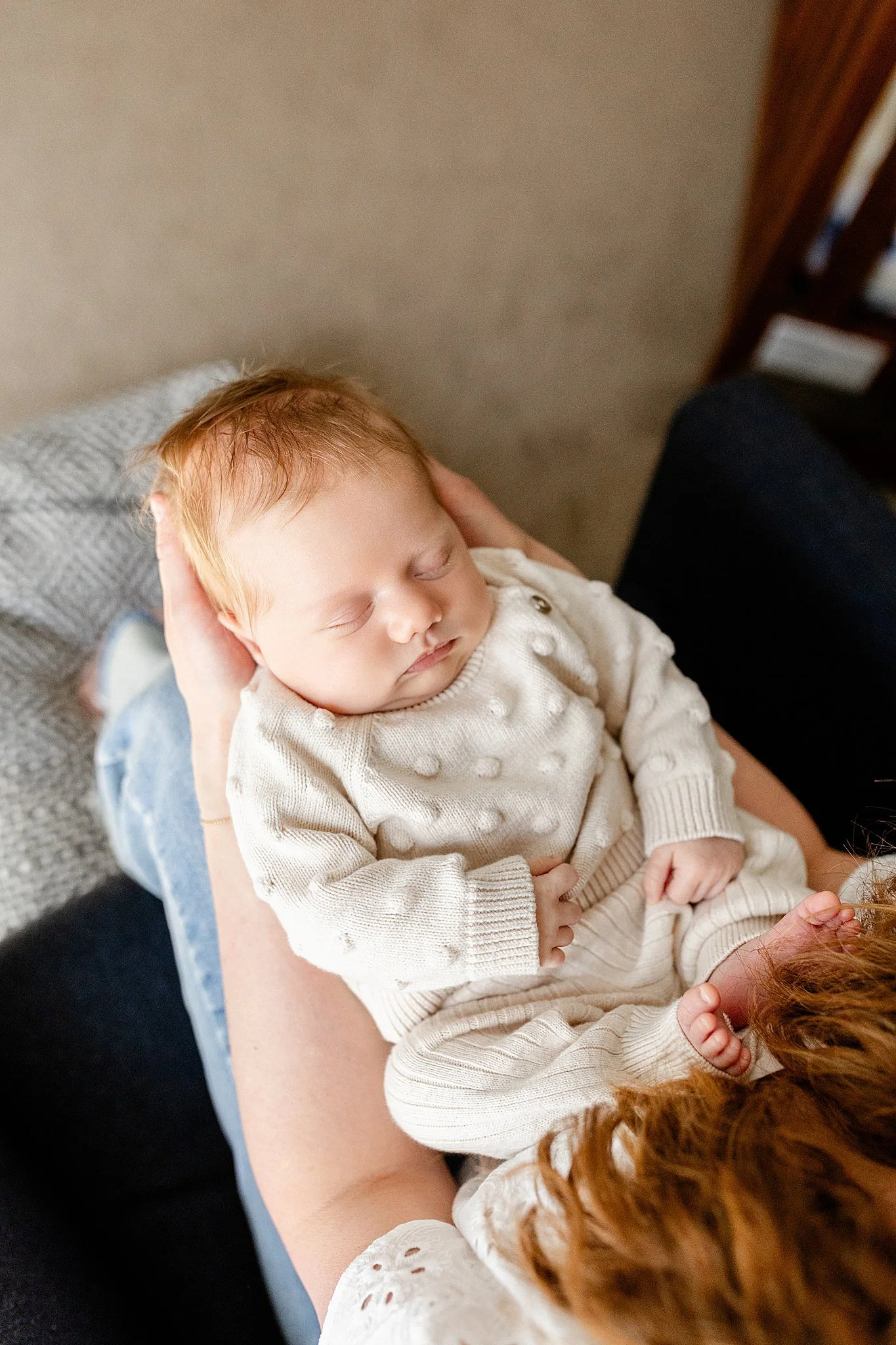 Close-up of a young child with red hair and closed eyes, resting on an adult's lap, with the adult holding the child's head gently with one hand. The child is wearing a cream-colored knitted sweater with textured dots with Ally and B Photography.