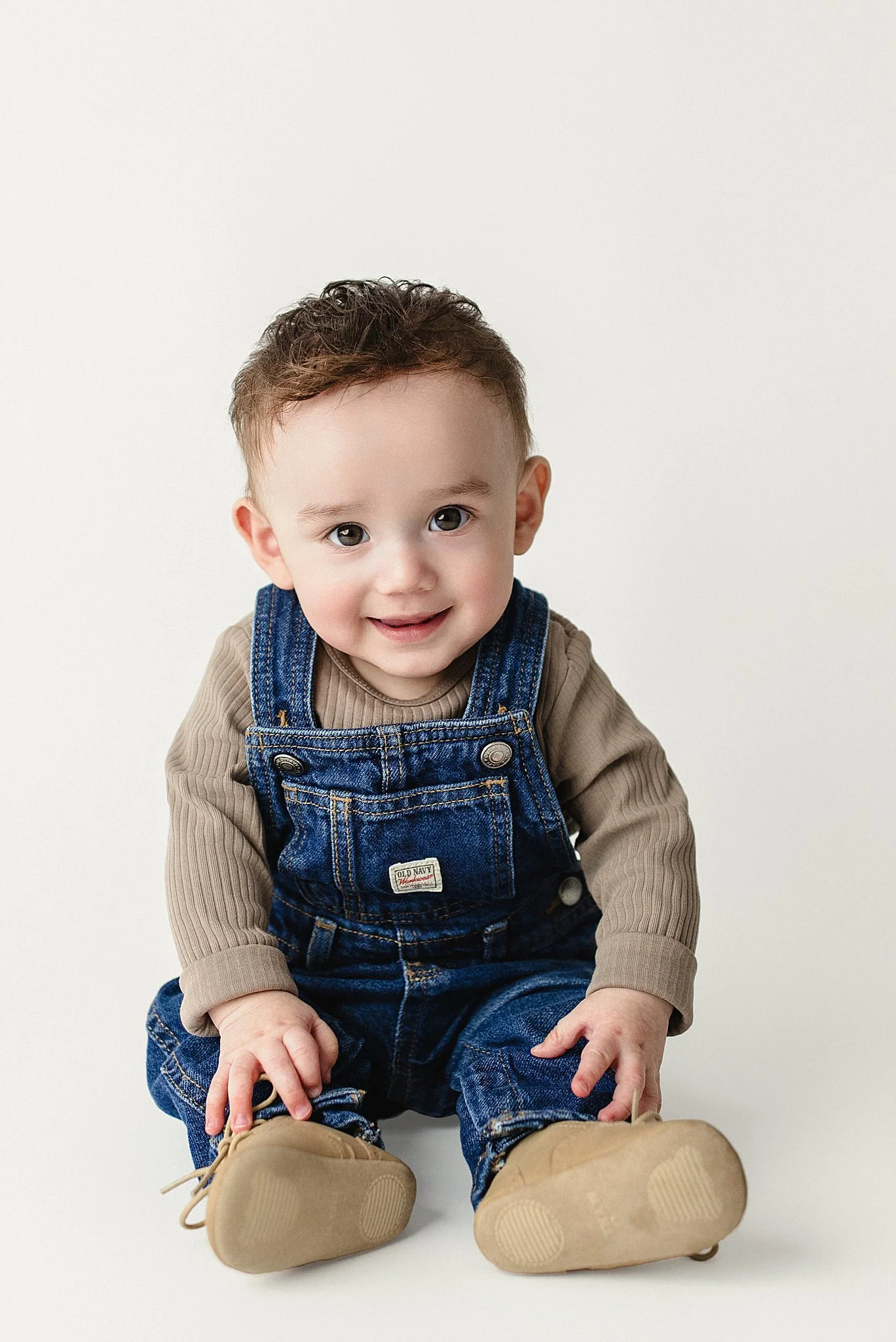 A smiling toddler sitting on the floor, wearing denim overalls, tan shoes, and a beige long-sleeve shirt with infant photographer Naperville.