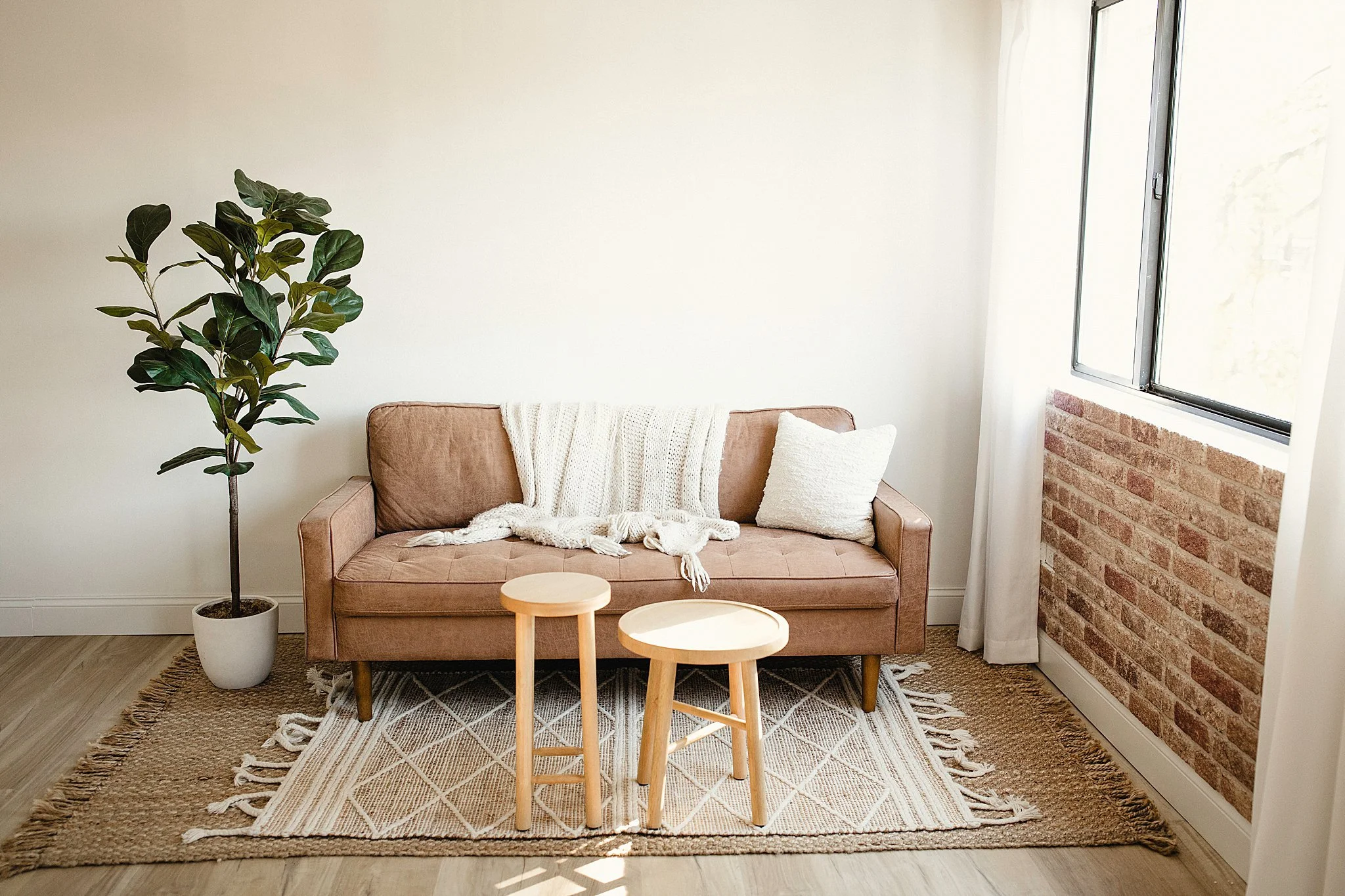 A cozy living room with a carmel sofa, white pillows, a throw blanket, two wooden stools, a plant in a white pot, a brick accent wall, and a large window with white curtains in a photo studio near me.