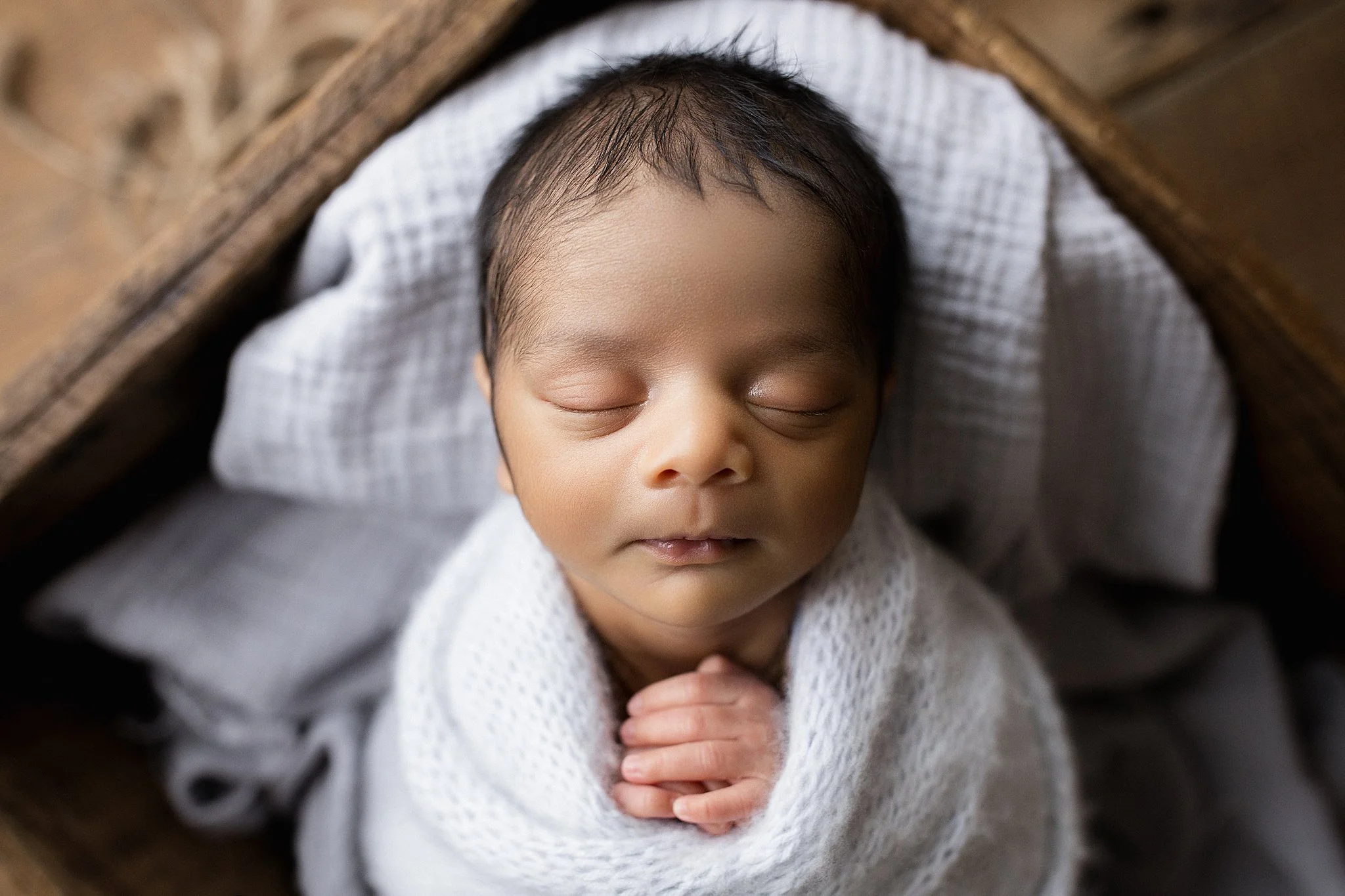 A sleeping newborn baby wrapped in a white blanket, lying on a checked pillow inside a wooden cradle during a newborn photoshoot.