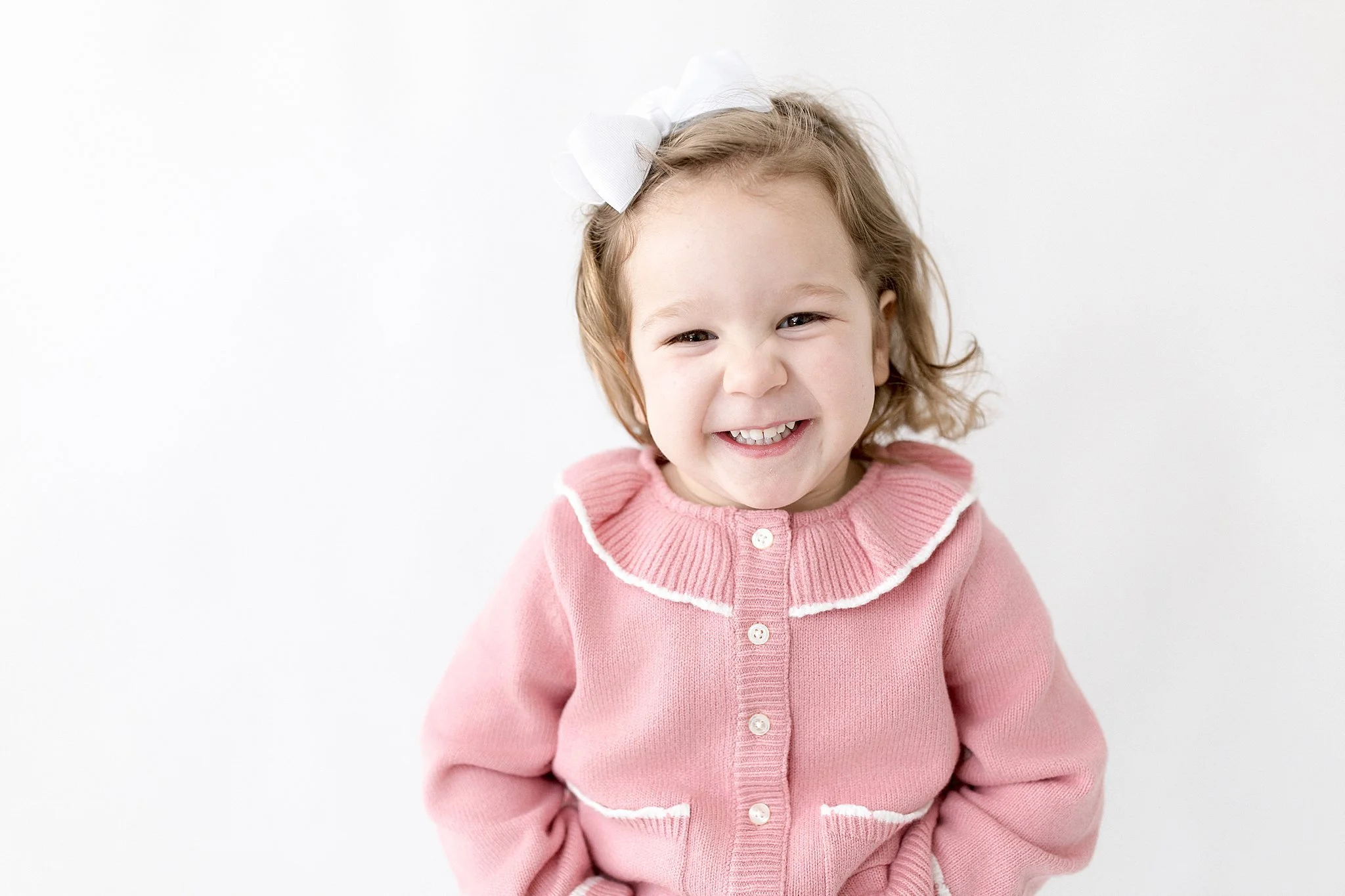 A young girl with light brown hair, wearing a pink cardigan with white trim and a white bow in her hair, smiling cheerfully with a kids portrait photographer near me.