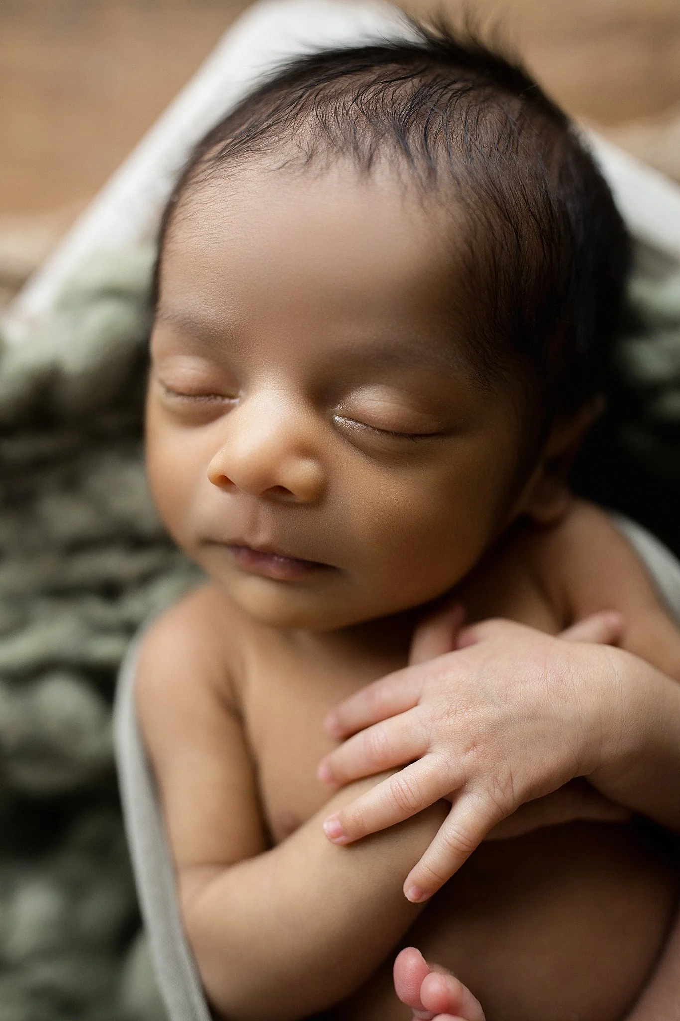 Close-up of a sleeping baby with dark hair, hands crossed on chest, resting on a soft blanket with the best newborn photographer in Naperville, IL.