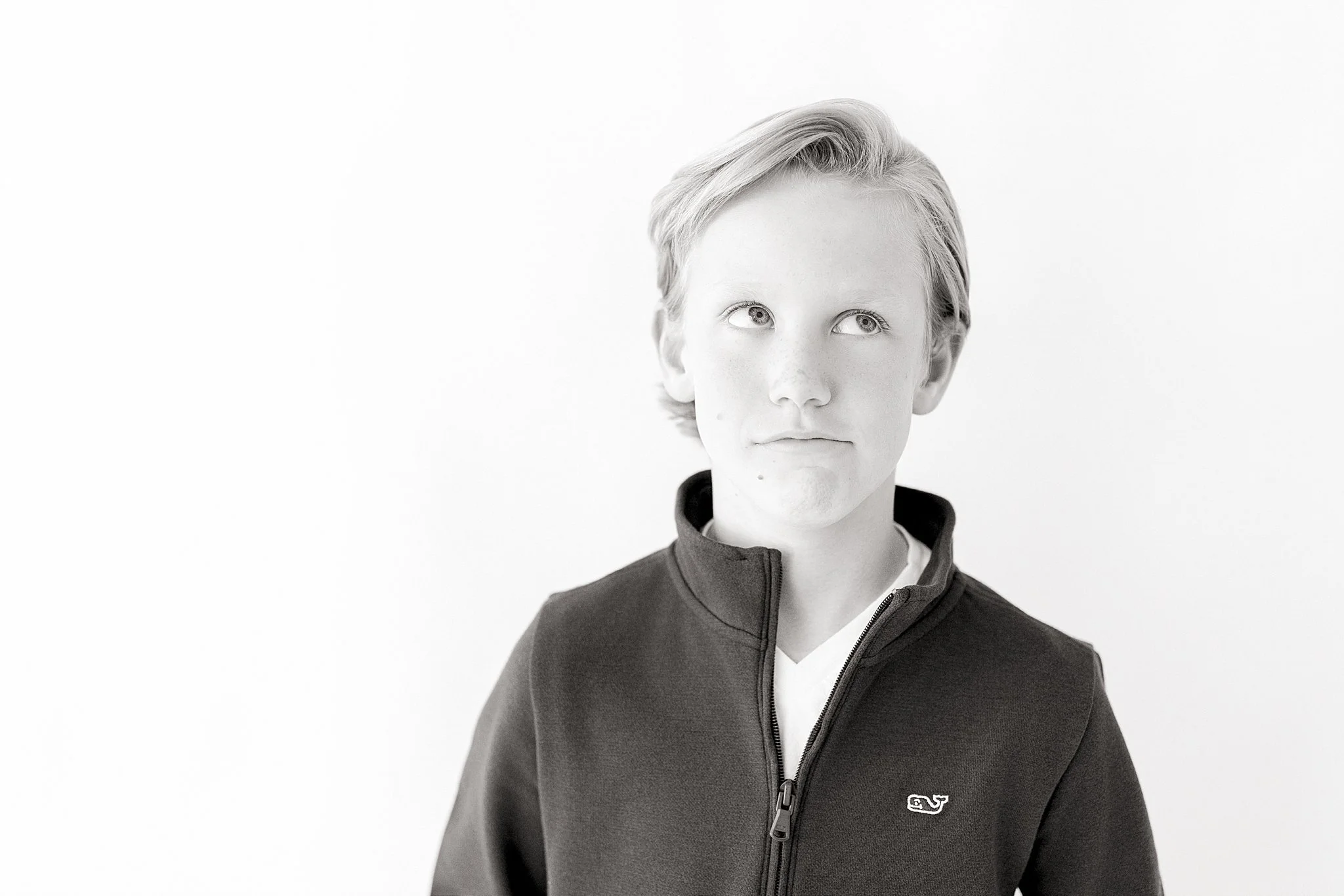 Black and white photo of a boy with light-colored hair, wearing a zip-up jacket with a small logo on the chest, looking to the side with a neutral expression with Ally and B Photography in Naperville, IL.