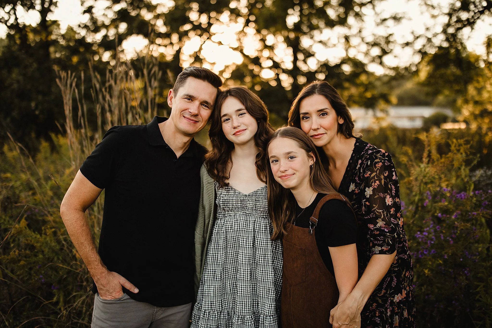 A family of four standing outdoors at sunset, smiling and posing together. The background features trees and purple flowers during a Naperville family photo session with Ally and B Photography.