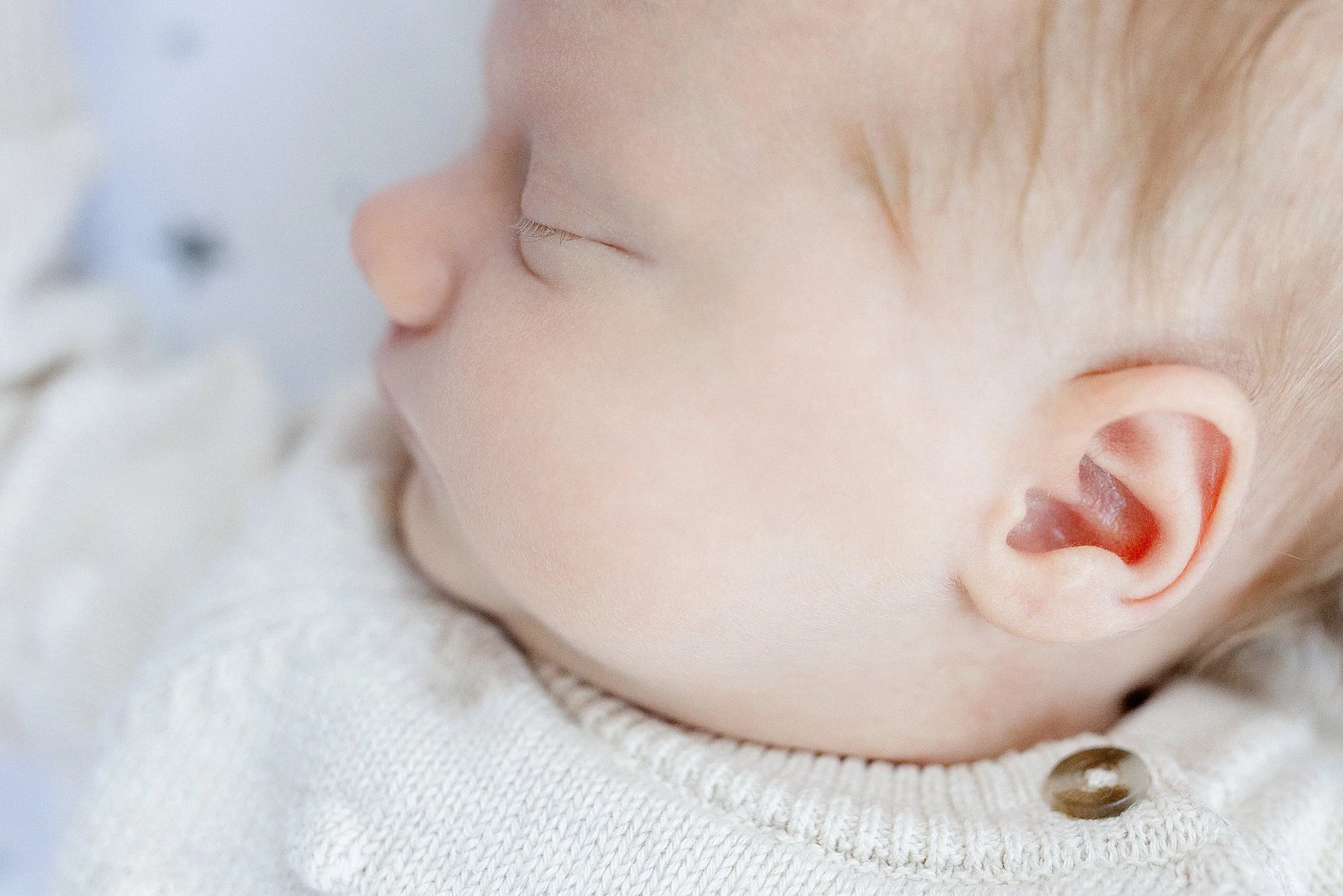 Close-up of a realistic baby doll's face showing closed eyes, a small nose, and a partially open mouth with a visible tongue, wearing a light-colored knit garment during in home lifestyle newborn photography near me.