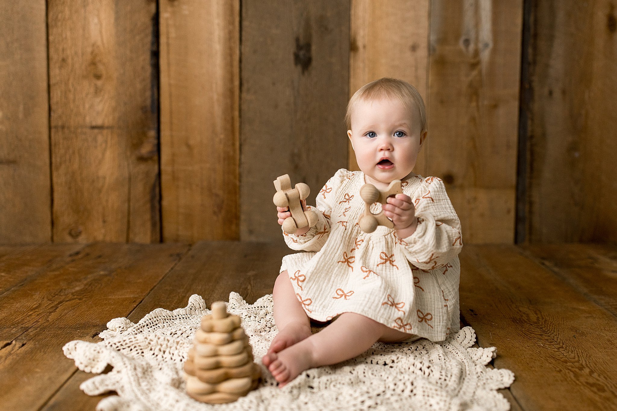 A baby girl with light skin and blonde hair sitting on a crocheted blanket on a wooden floor, holding a wooden toy car and looking at the camera, with a wooden wall in the background during a baby photoshoot in downtown Naperville, IL.