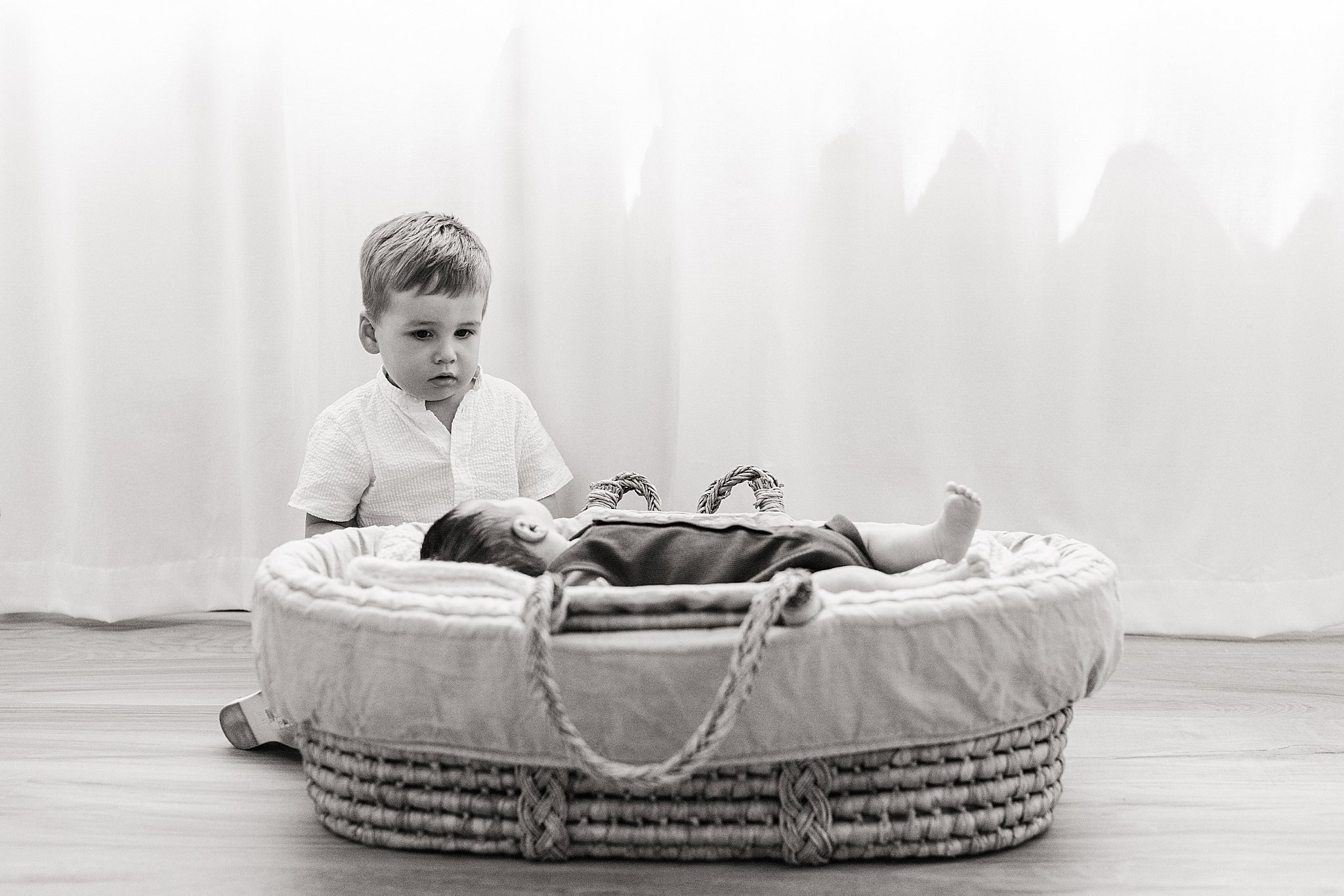 A young boy looking at a baby lying in a woven basket on a wooden floor during a lifestyle newborn photo session in downtown Naperville, IL.